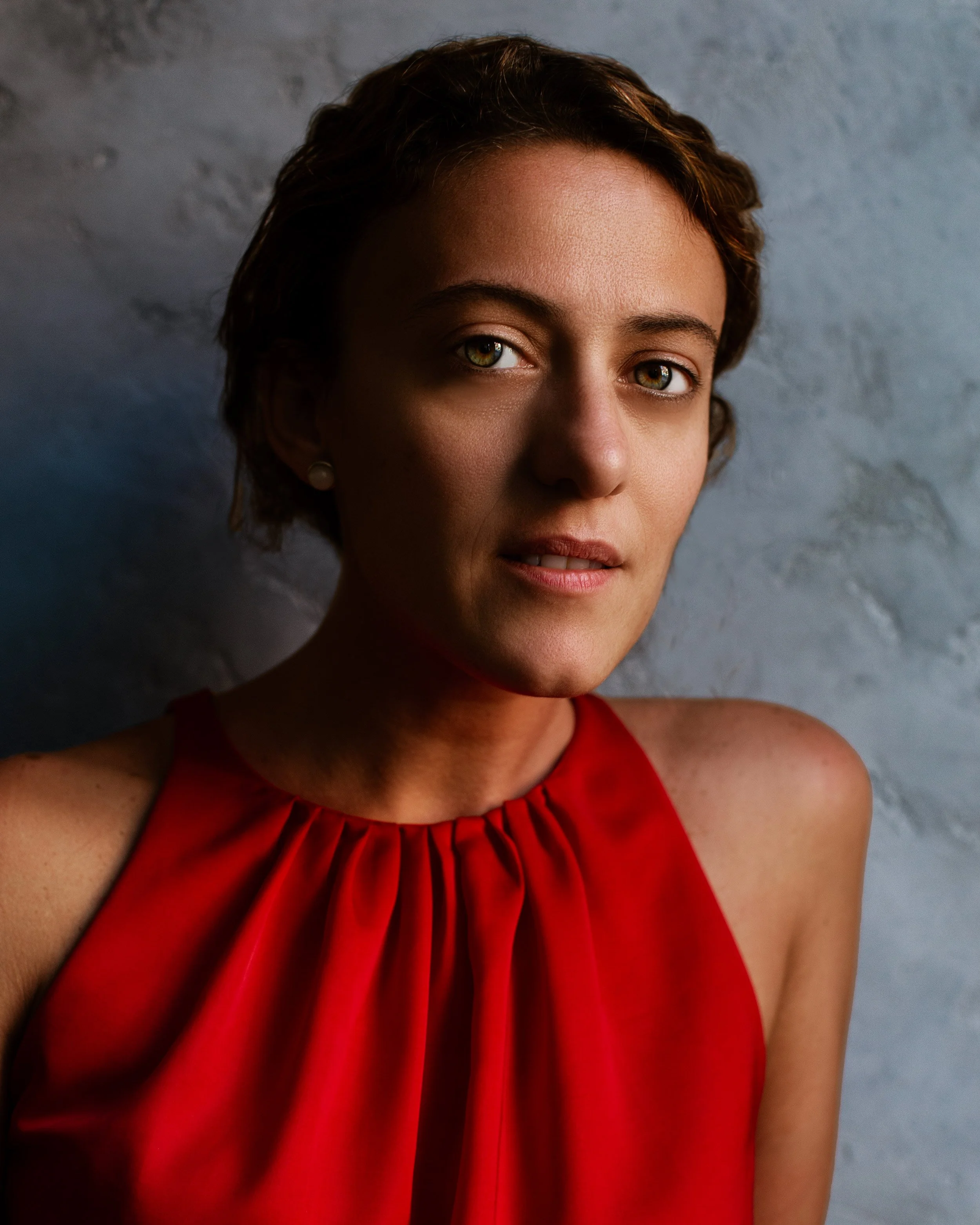 Close-up of a woman wearing a red top with pleated neckline, smiling slightly, against a textured gray background.