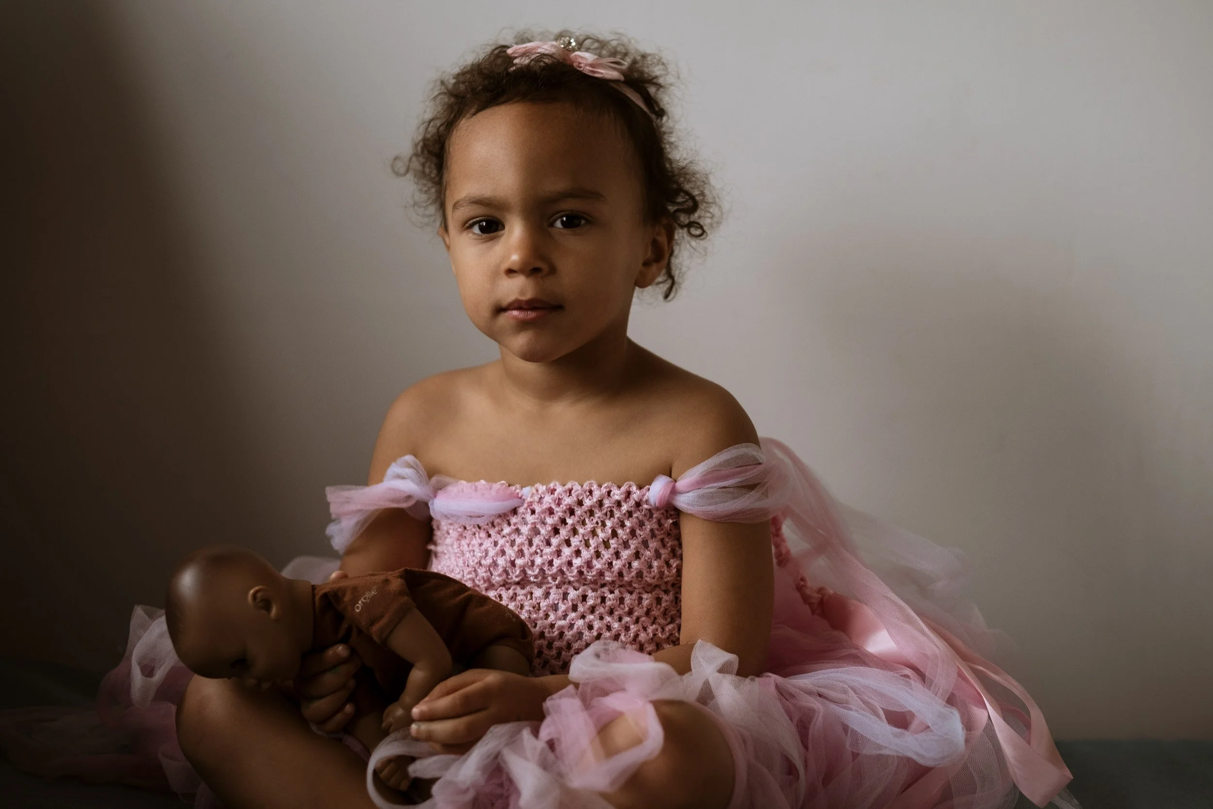 Young girl with curly hair wearing a pink dress and headband, holding a baby doll, sitting against a plain wall.