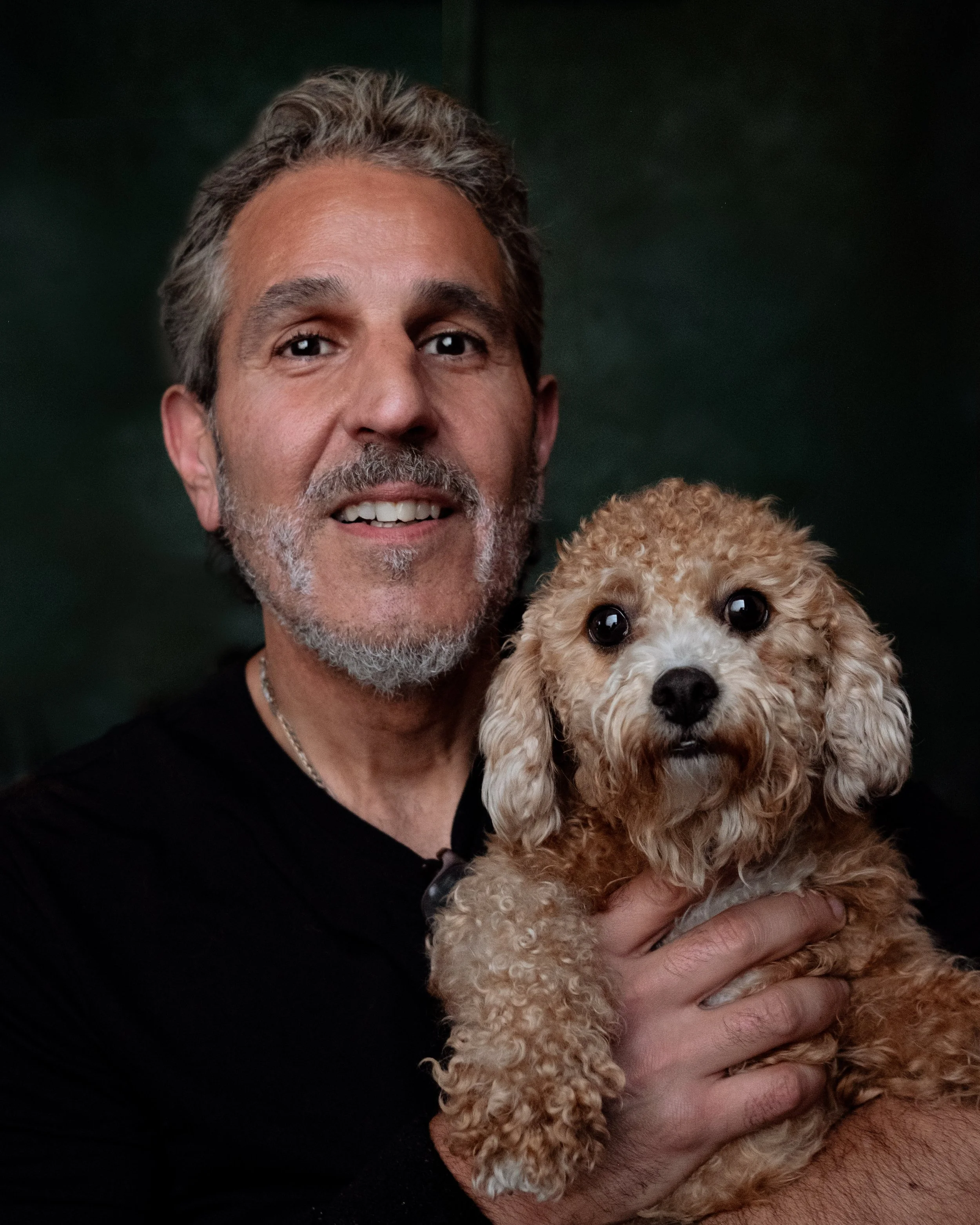 A man with gray hair and a beard holding a small, curly-haired, light brown poodle.