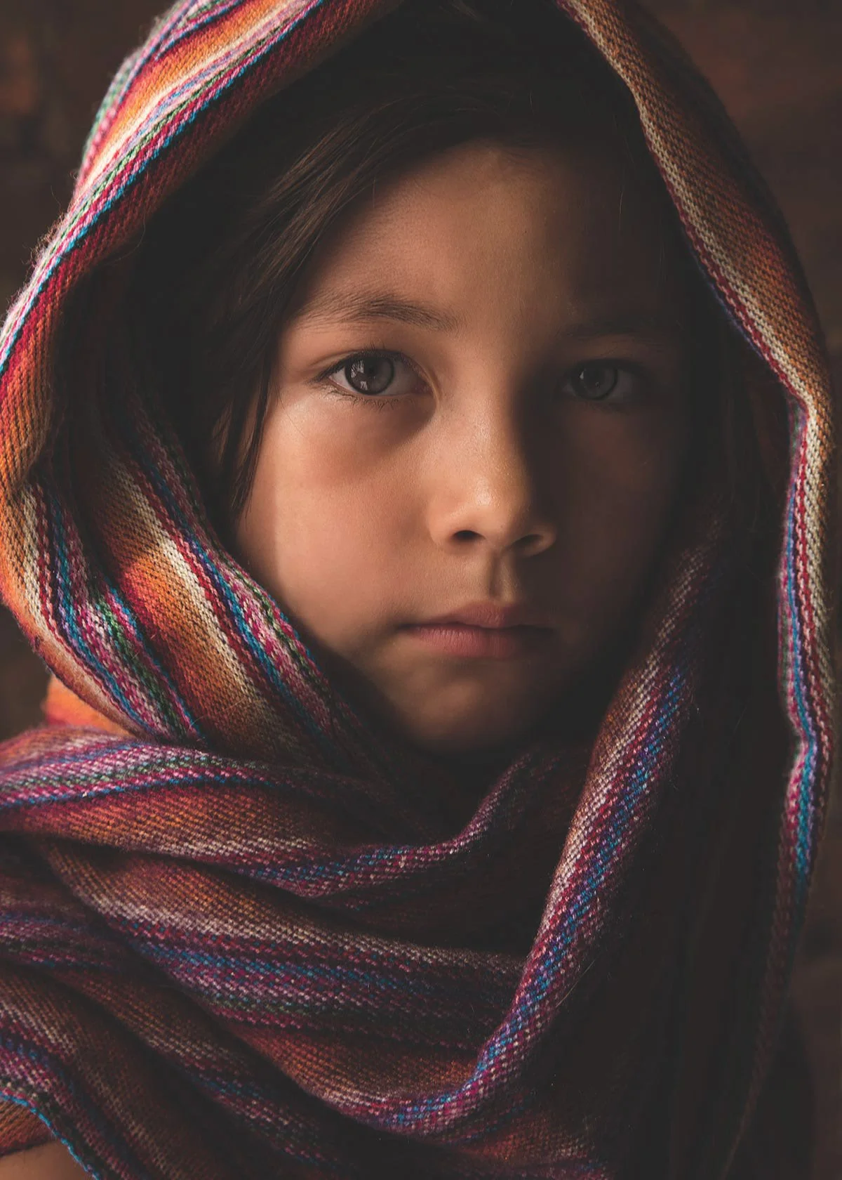 Close-up portrait of a young girl with light skin and dark hair, wrapped in a colorful, patterned scarf over her head, with a neutral expression and light-colored eyes.