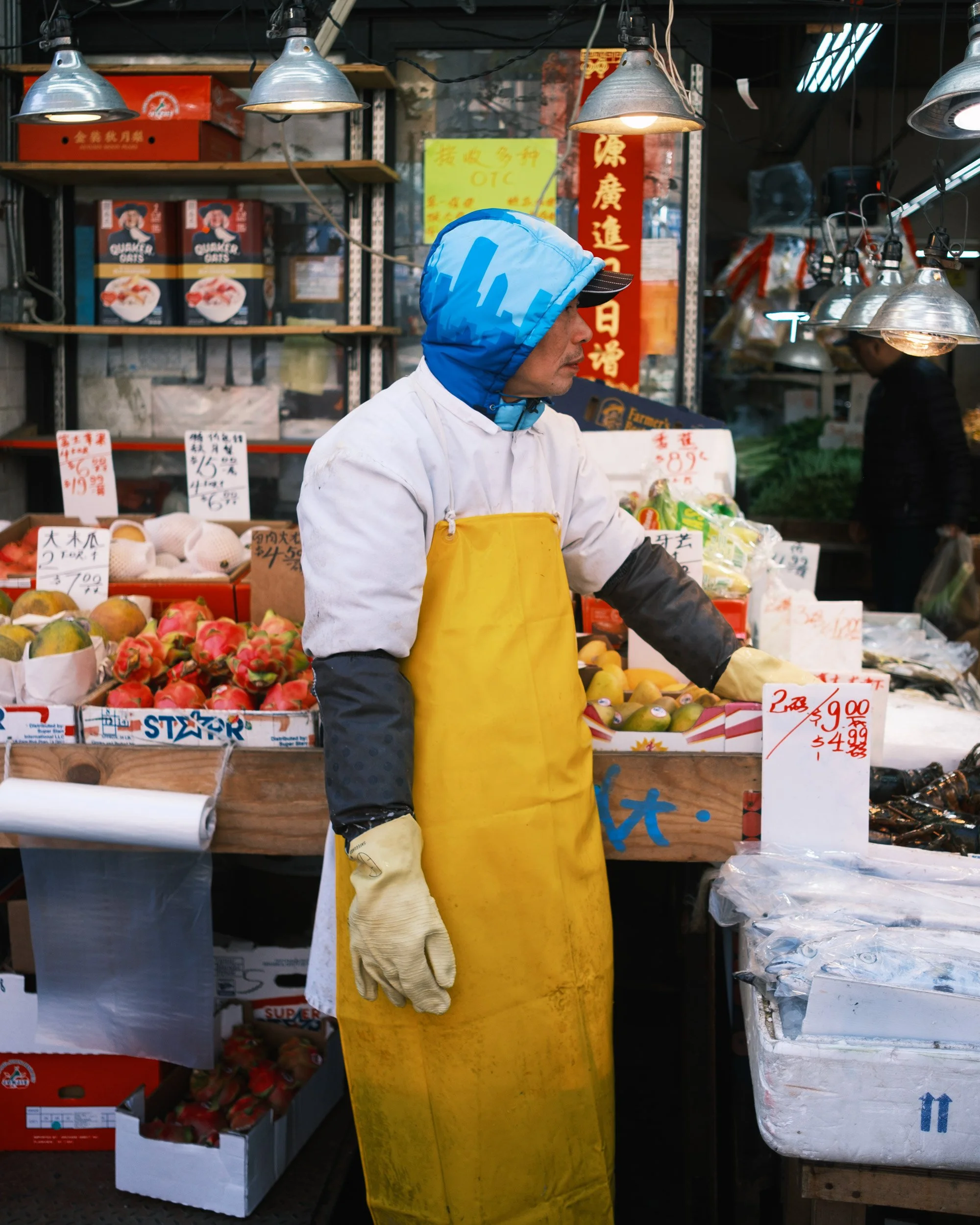 A man wearing a yellow apron and a blue hooded jacket with cityscape design is shopping at an indoor market stall. The stall displays various fresh fruits including apples, peaches, and kiwis, with handwritten price tags. Overhead, metallic lamps ill