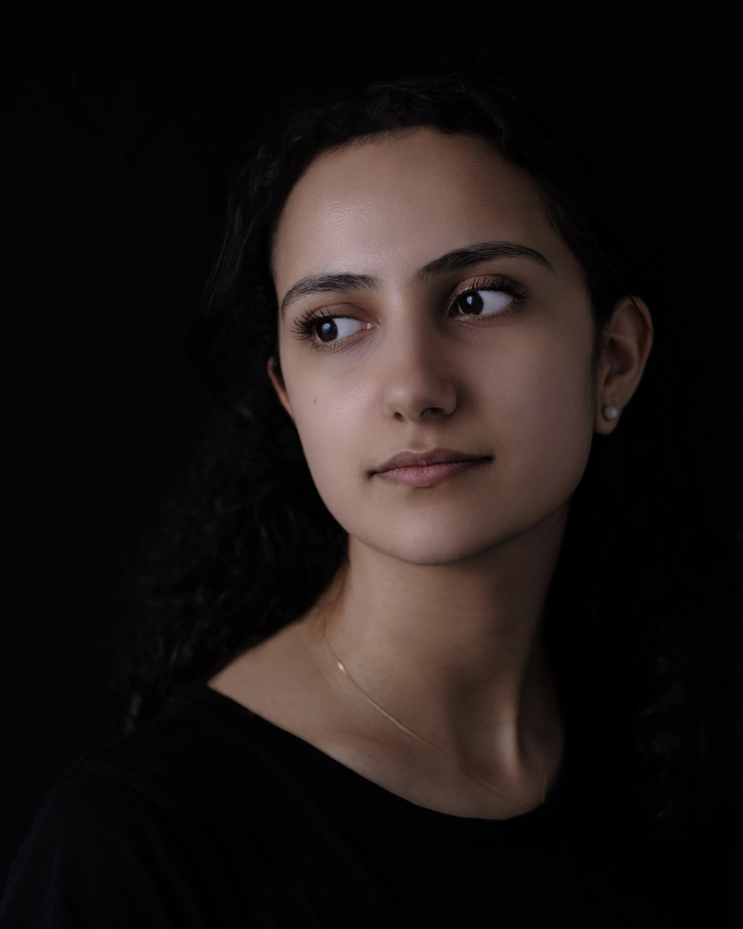 Close-up portrait of a young woman with dark, wavy hair, light skin, wearing pearl earrings, a thin gold necklace, and a black top, against a black background.