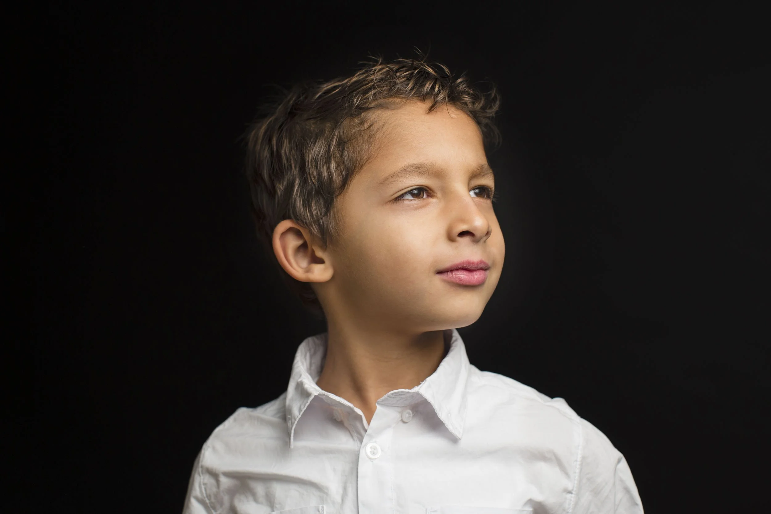 Young boy with brown curly hair and light skin, wearing a white collared shirt, looking to the side against a black background.