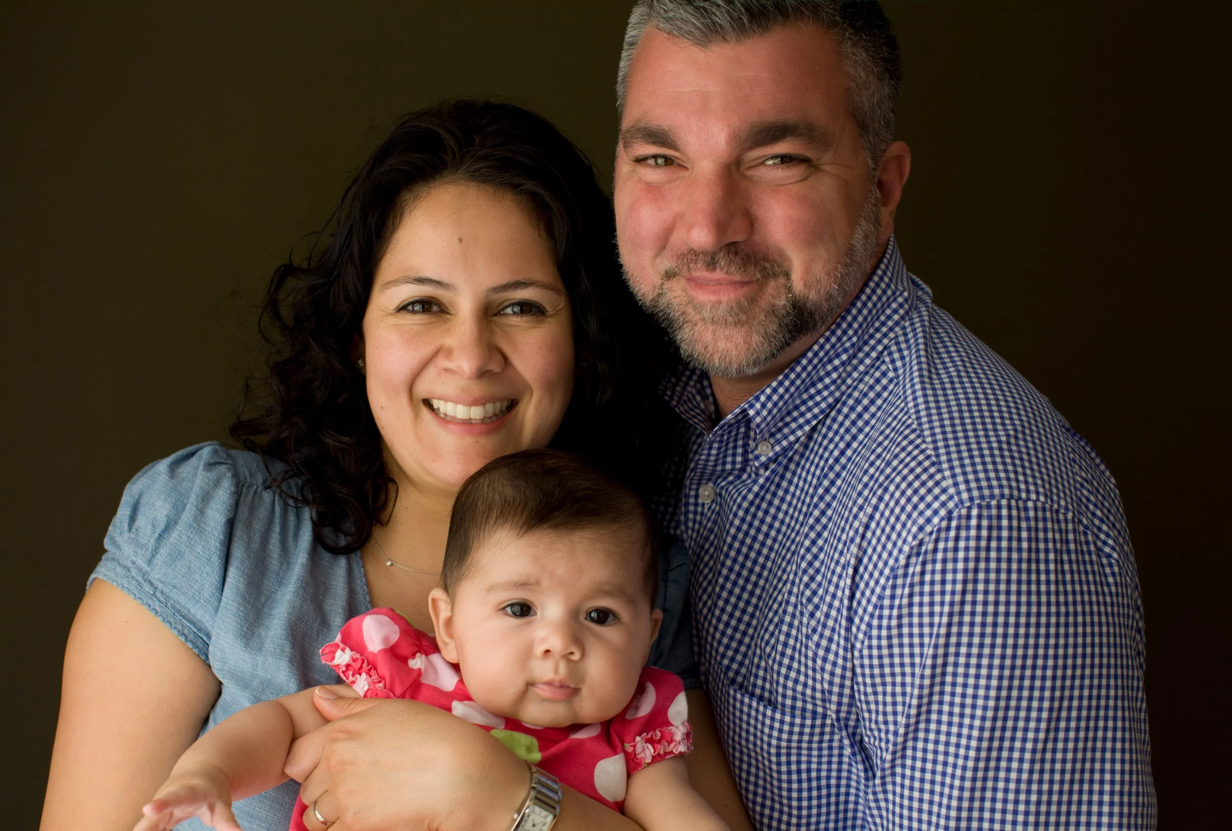 A happy, diverse family portrait featuring a woman with dark, curly hair, a man with short gray hair and beard, and a young child with light skin and dark hair, against a dark background.