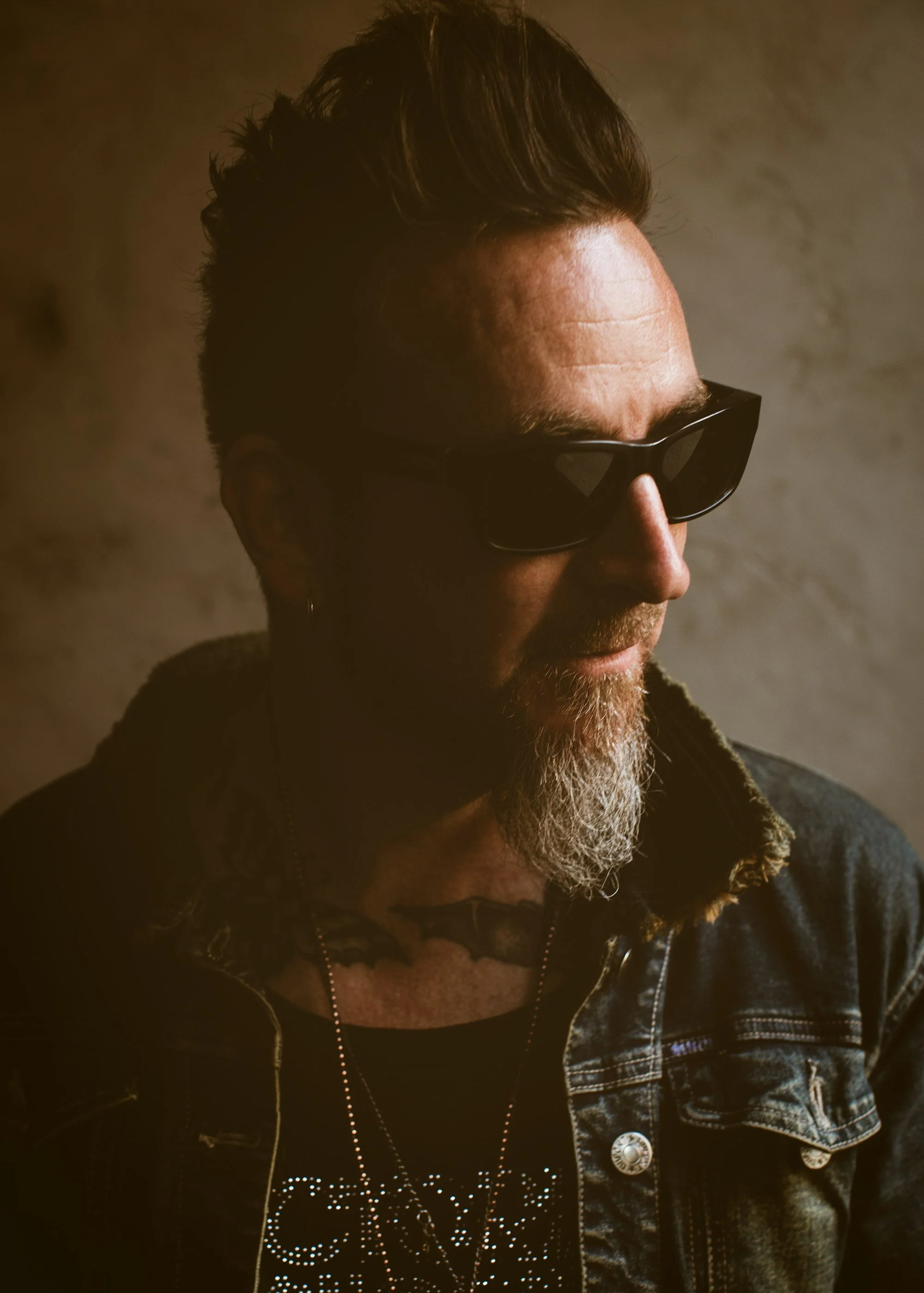 A man with a beard and wearing sunglasses, denim jacket, and a hoodie, looking to the side against a plain background.
