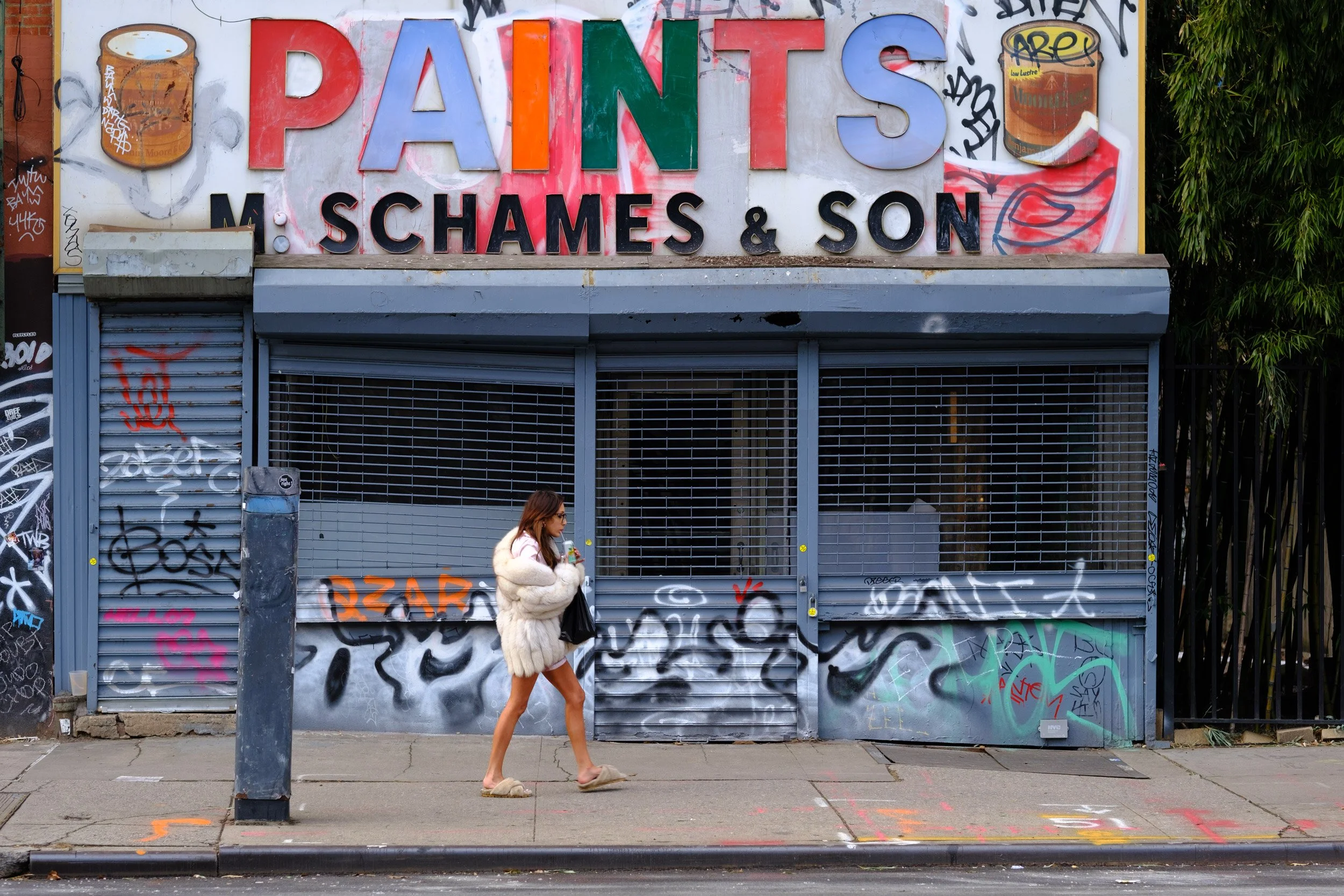 A woman with long brown hair wearing a fluffy light-colored coat, sunglasses, and tan slippers walking past a graffiti-covered metal storefront shutter and wall, with large colorful letters spelling 'PAINTS' and smaller letters 'S CHAMES & SON' in bl