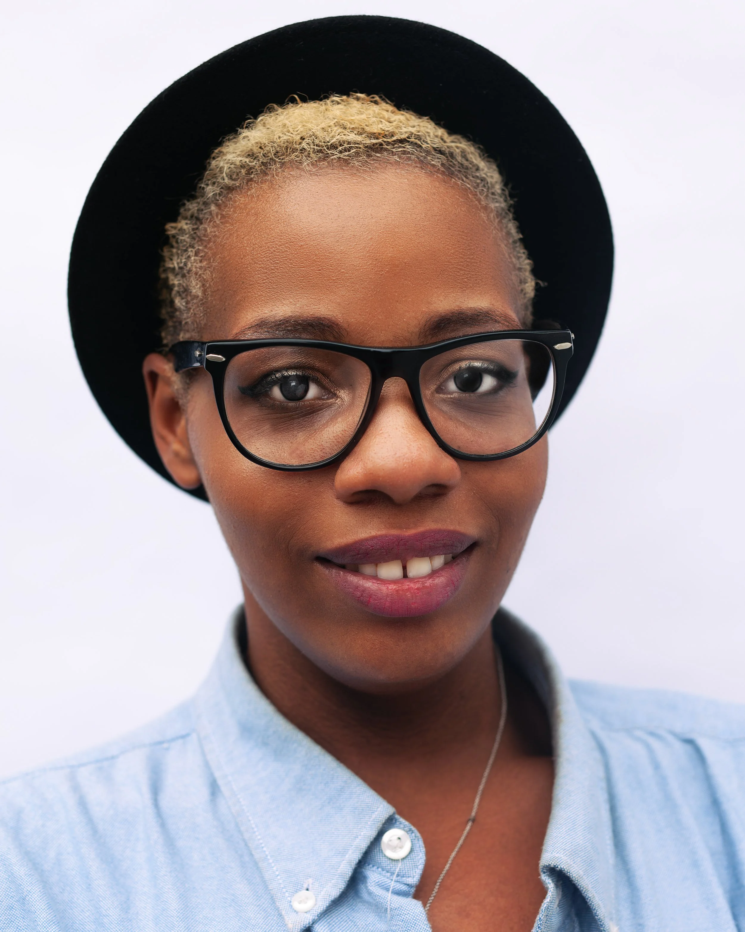 Close-up of a woman wearing a black hat, glasses, and a light blue shirt, smiling with part of her face visible.