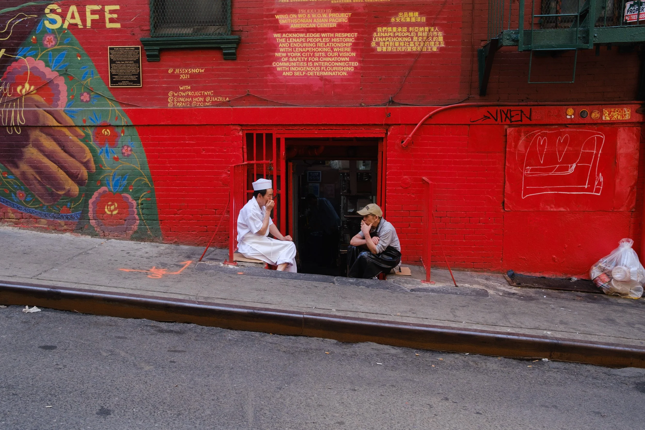 A person in a white chef's uniform and hat sitting on a step outside a red brick building with colorful street art and yellow text, talking to another person wearing glasses and a beige cap. There is graffiti on the wall and trash bags on the sidewal