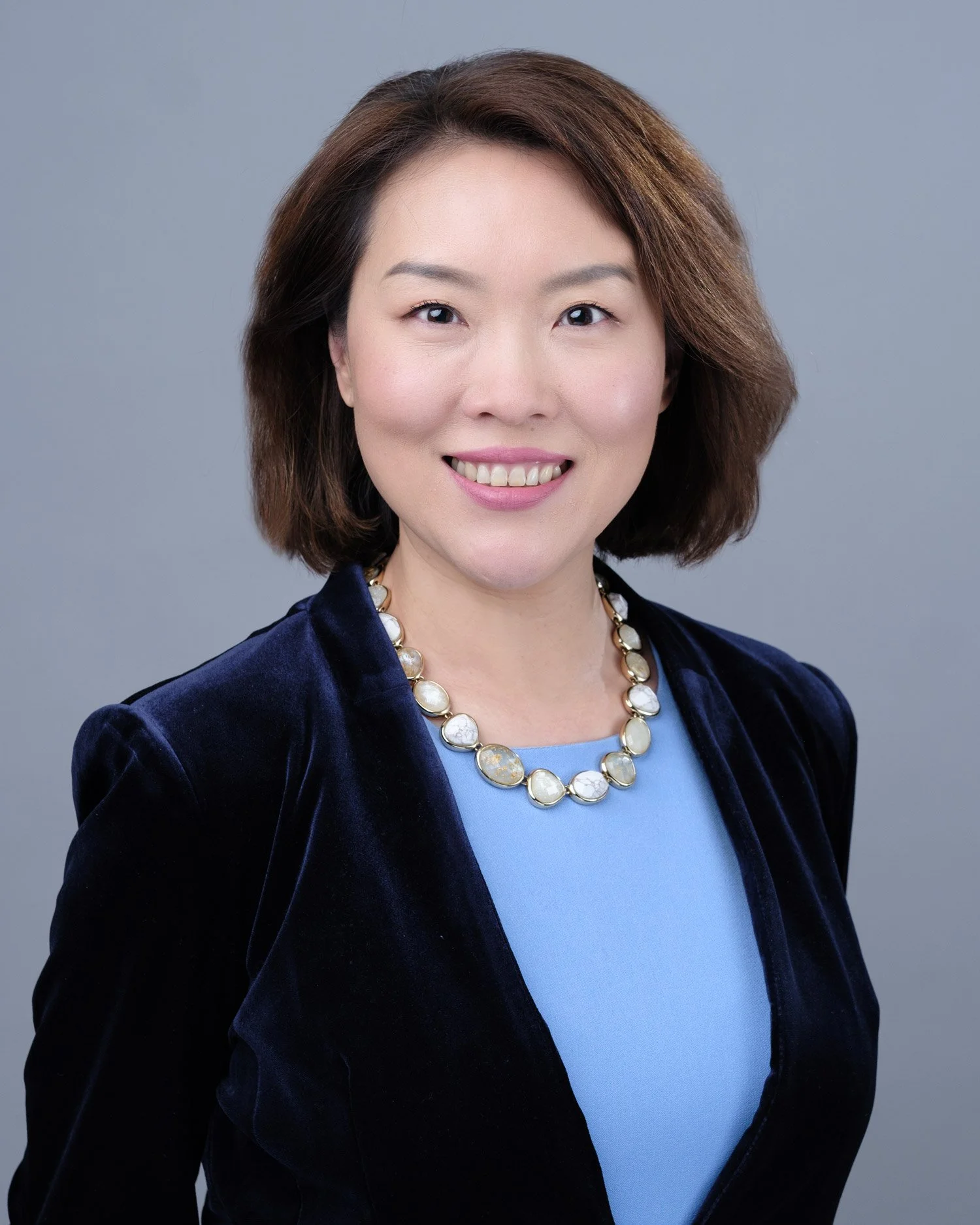 Headshot of an Asian woman with shoulder-length brown hair, wearing a dark blue velvet blazer, a light blue top, and a pearl necklace, smiling against a plain gray background.