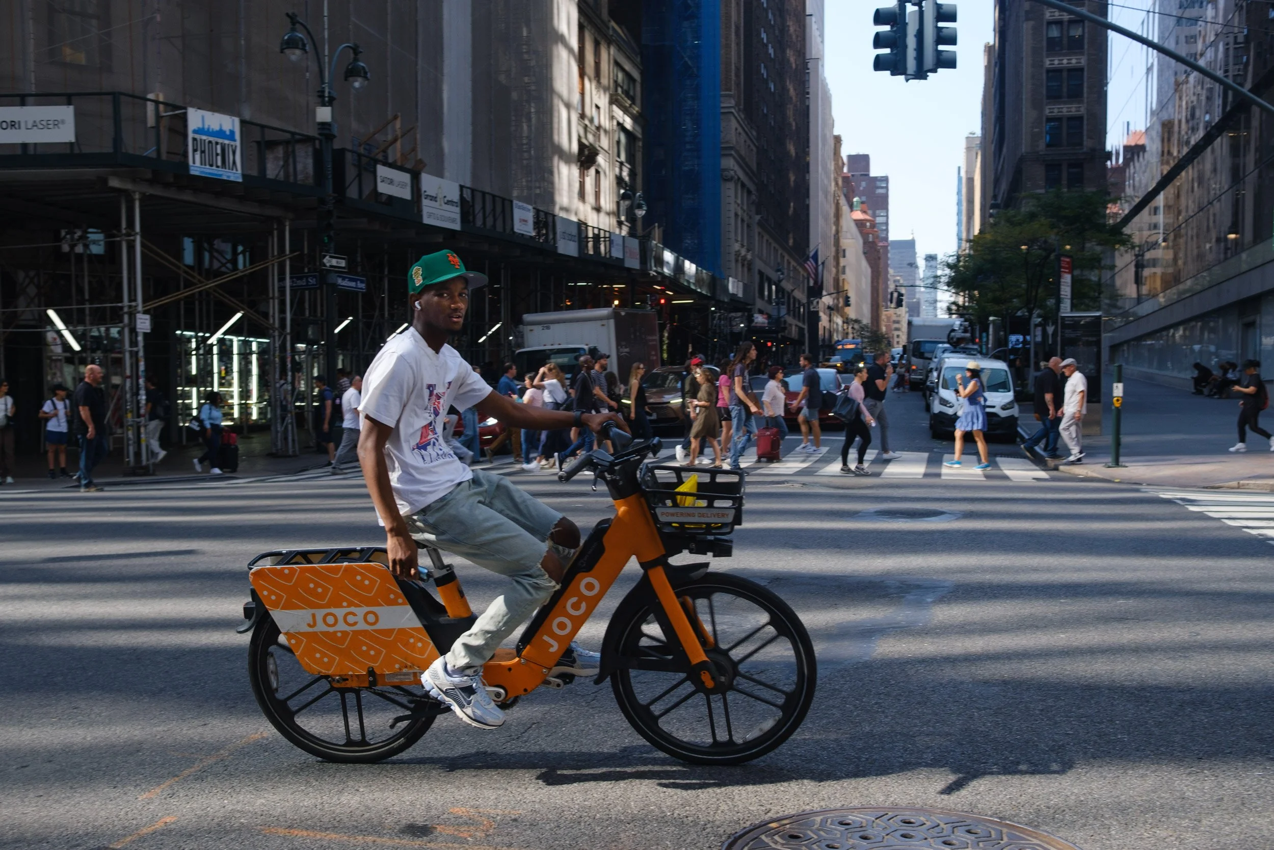 A man riding a yellow Joco bicycle across a busy city street with pedestrians walking on the crosswalk and tall buildings in the background.