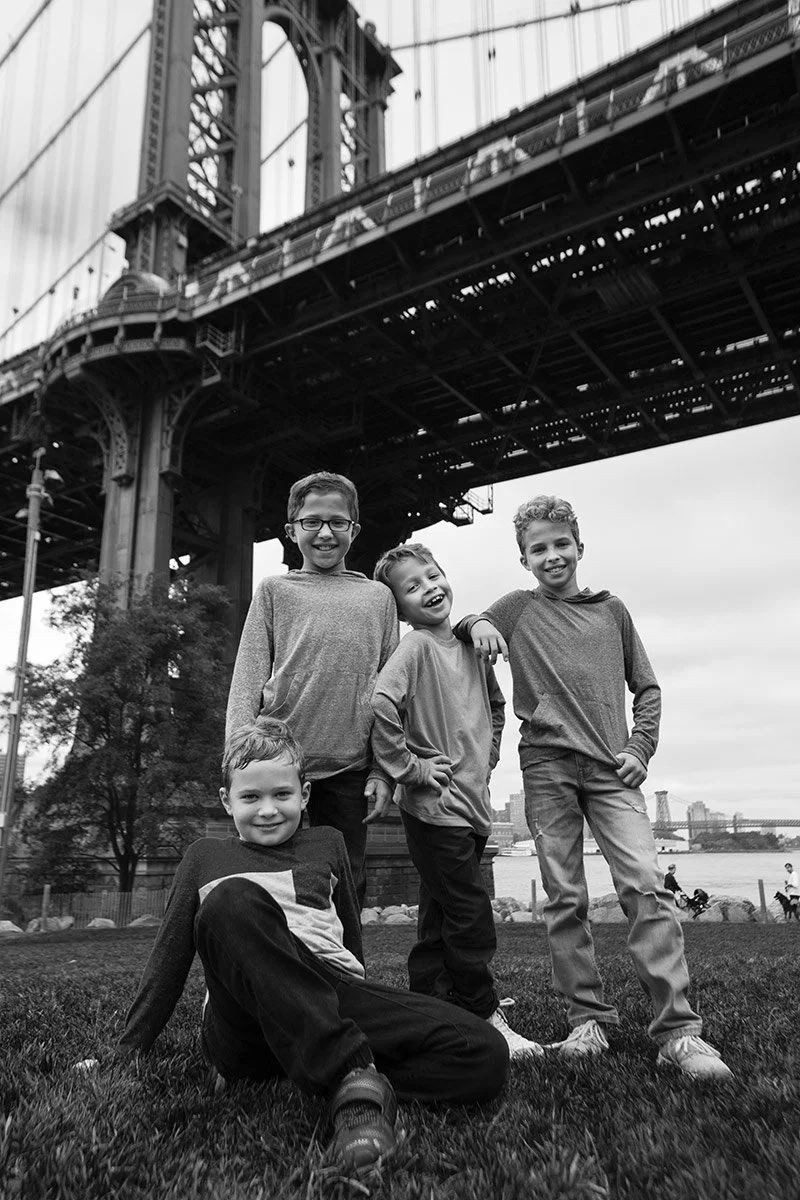 Four boys posing smiles under a bridge near a river, with a city skyline in the background.