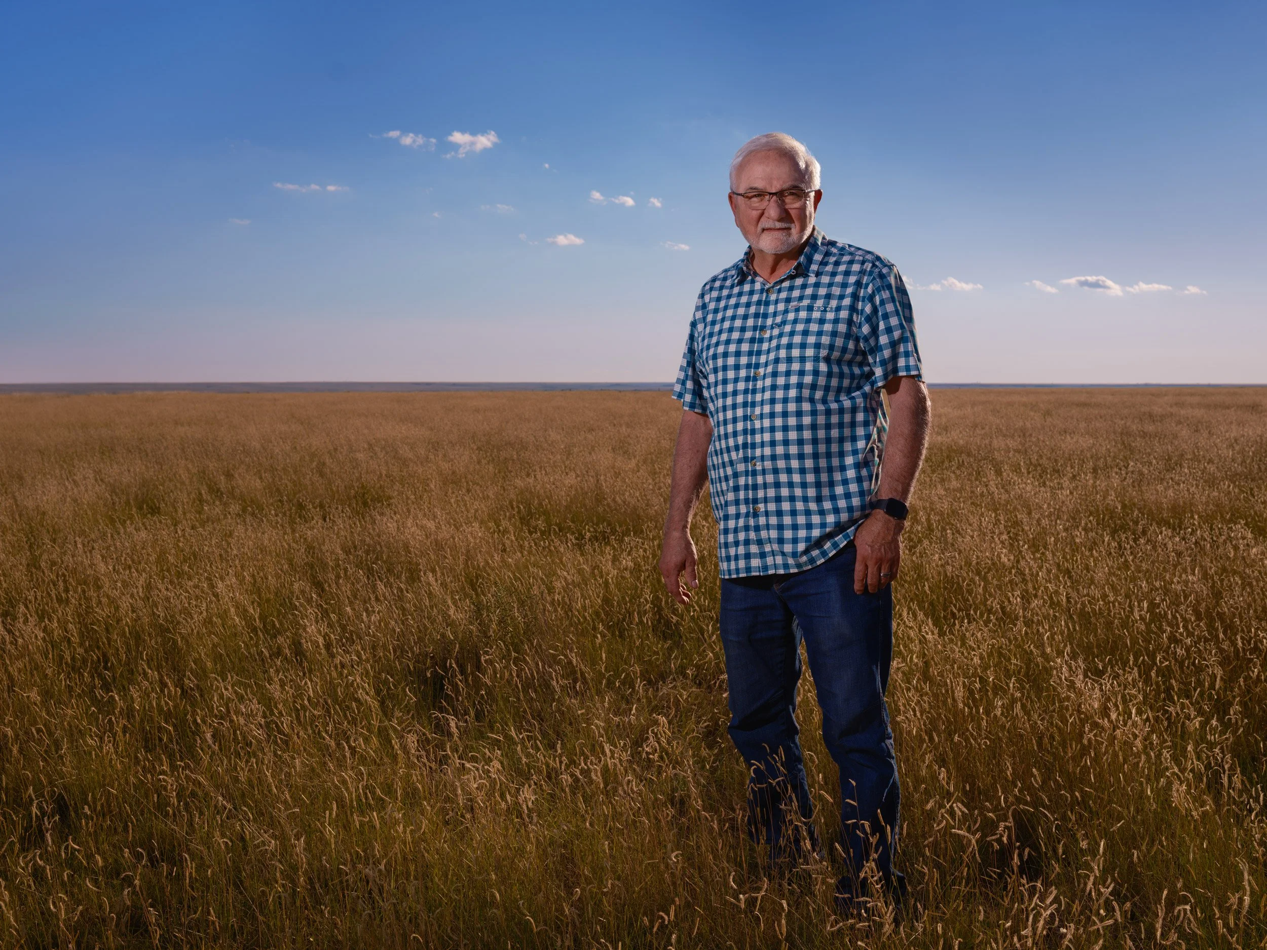 Older man with glasses and gray hair, wearing a blue checkered shirt and jeans, standing in a field of tall grass under a clear blue sky.