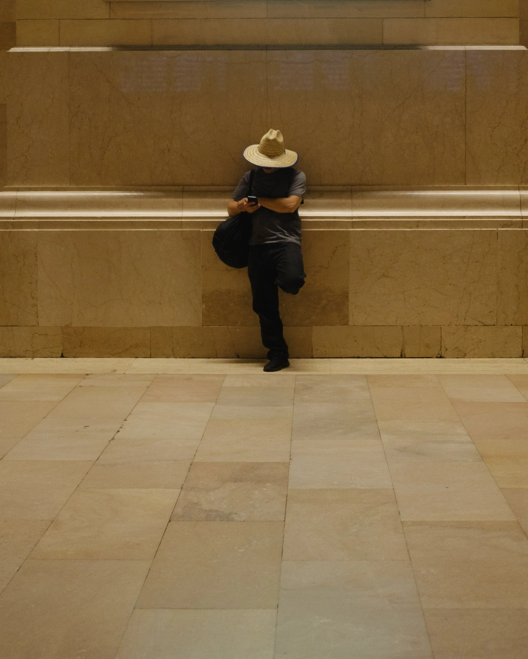 A person leaning against a marble wall, wearing a wide-brimmed straw hat, dark gray t-shirt, dark pants, and holding a smartphone, with a dark bag over their shoulder.