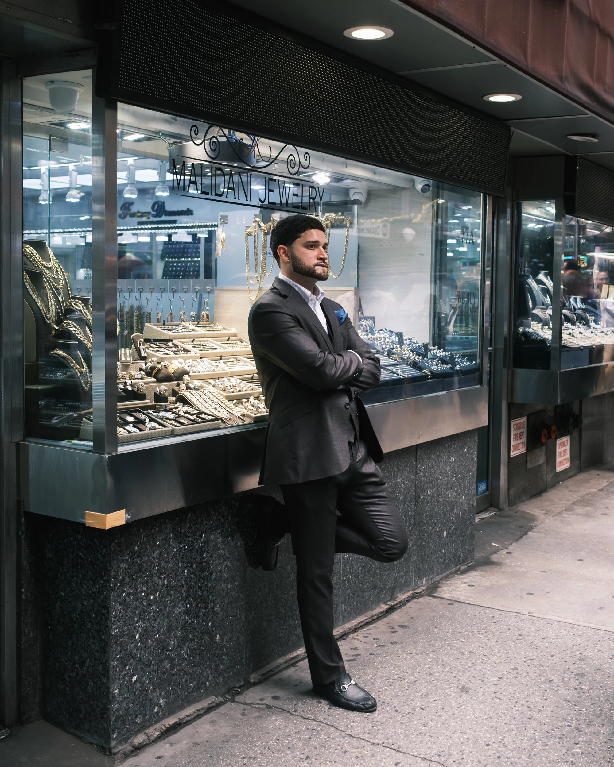 A man in a suit leaning against a jewelry store window with his arms crossed and one foot up on the wall outside the store.
