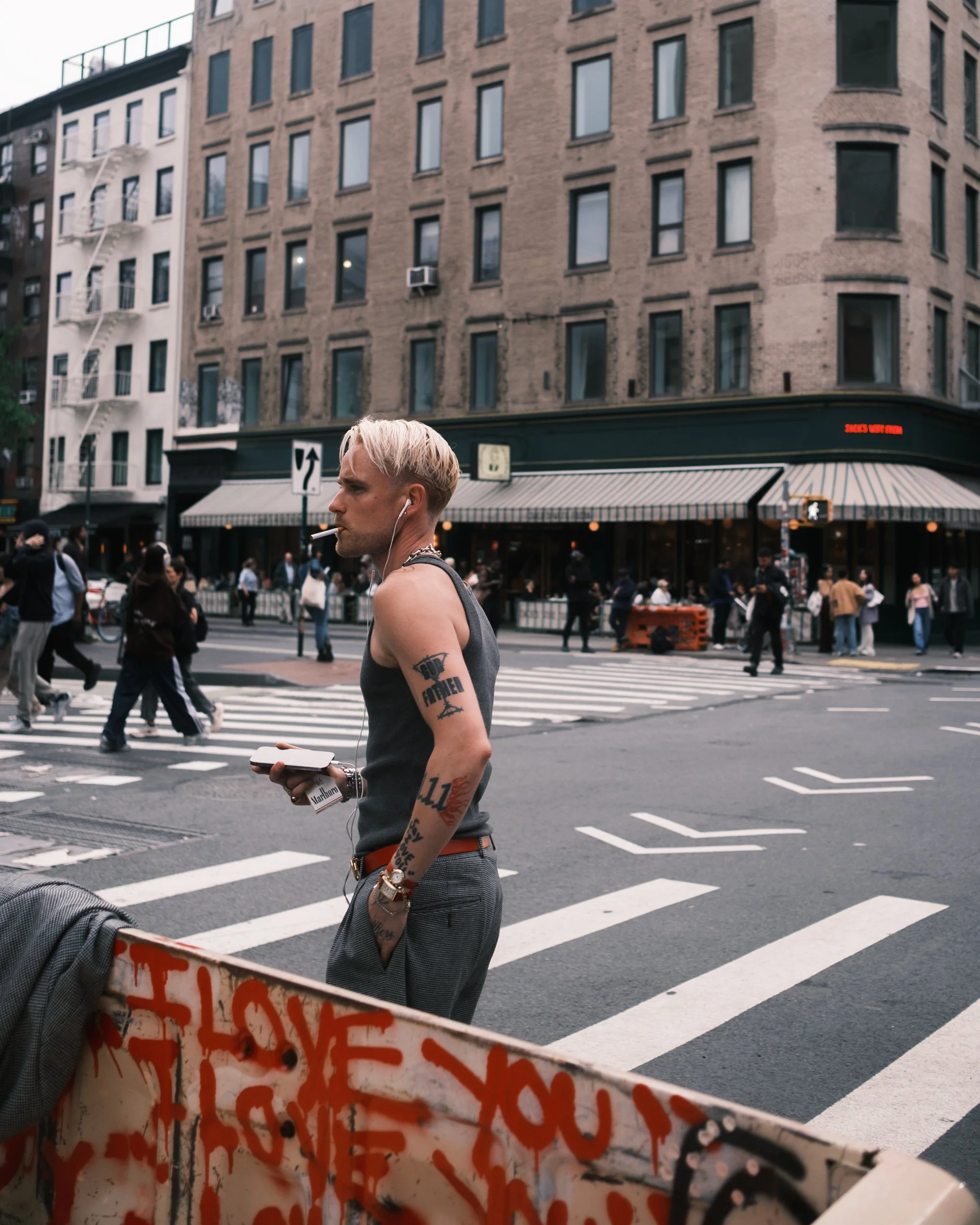A young man with blonde hair and tattoos on his arms, smoking a cigarette, walking on a busy city street with a crowd of people crossing the intersection. The background shows a large multi-story building with brick and white walls, several windows, 