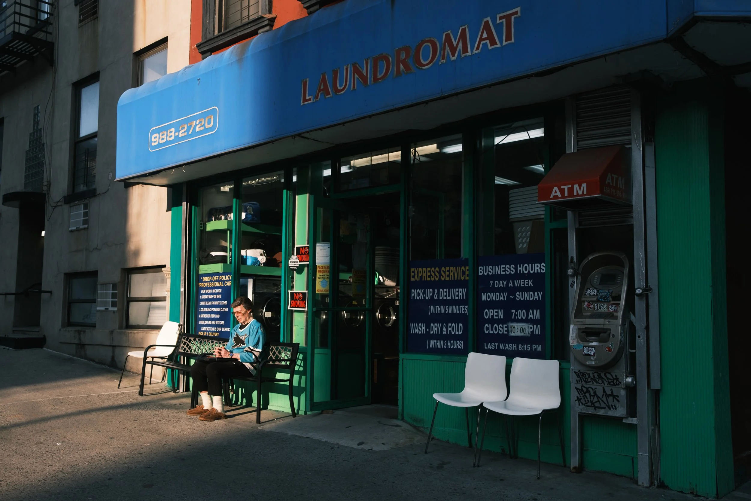 A laundromat with green exterior and blue awning, showing the word 'LAUNDROMAT' and the phone number 988-2720. There are service signs and an ATM outside, and a person sitting on a bench in front of it. Two empty white chairs are next to graffiti-cov
