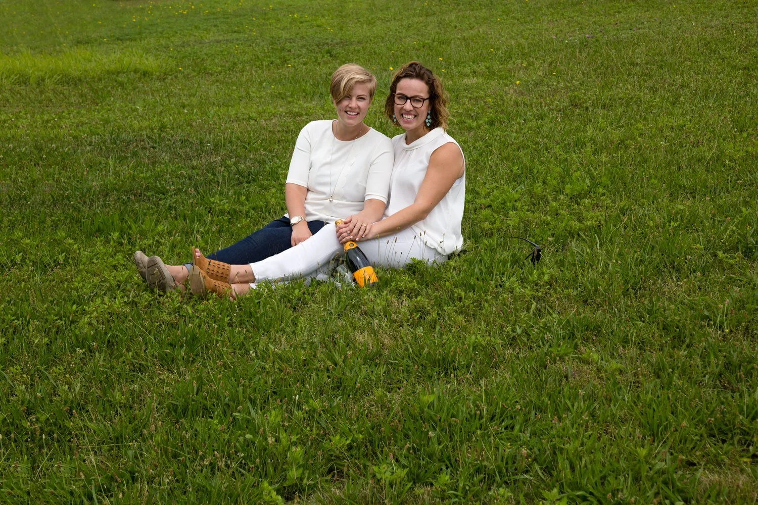 Two women sitting on green grass in a park, smiling and holding hands, with a small orange and black wine bottle placed on the ground.