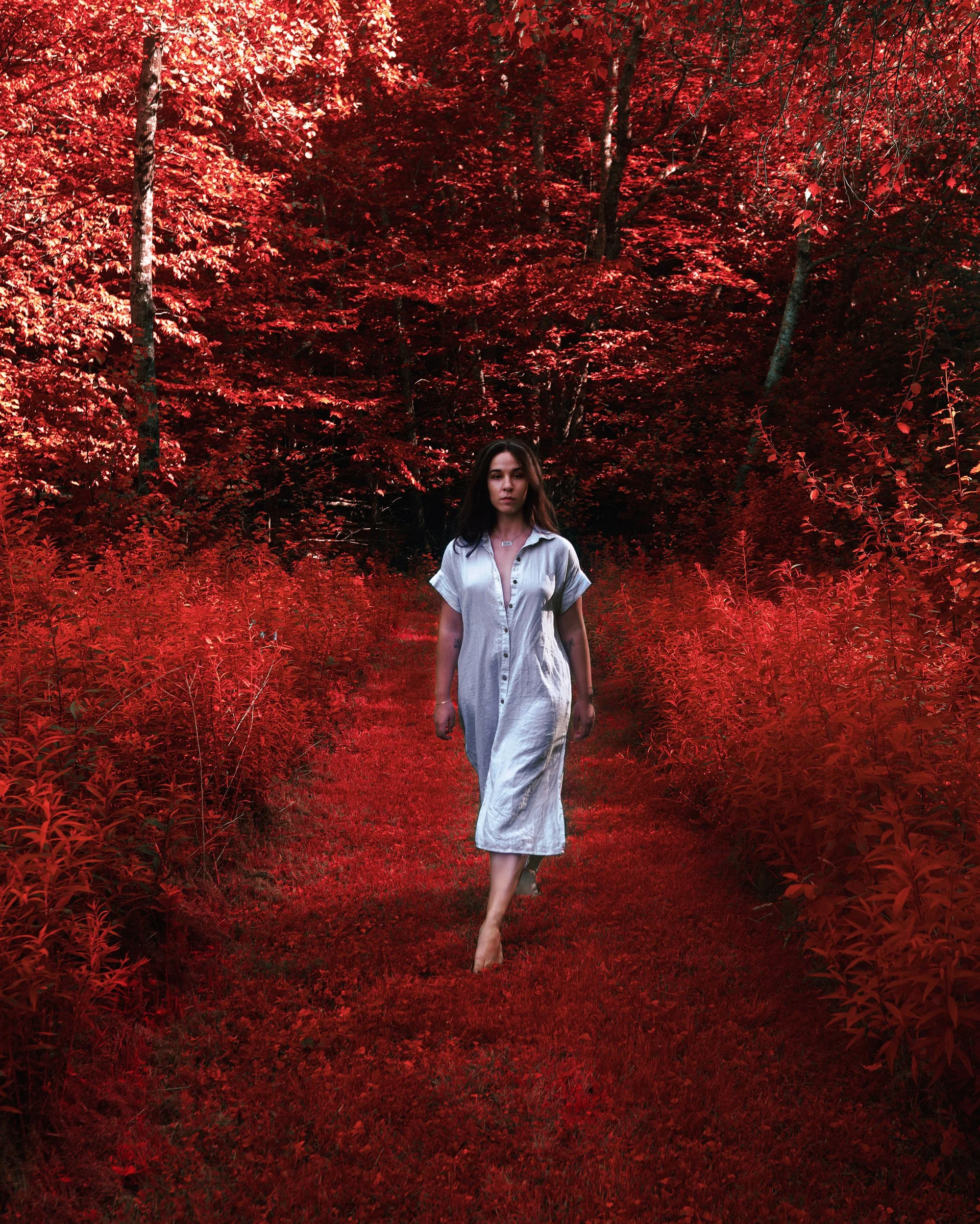 A young woman walking barefoot on a red forest trail surrounded by vibrant red foliage with dense trees in the background.