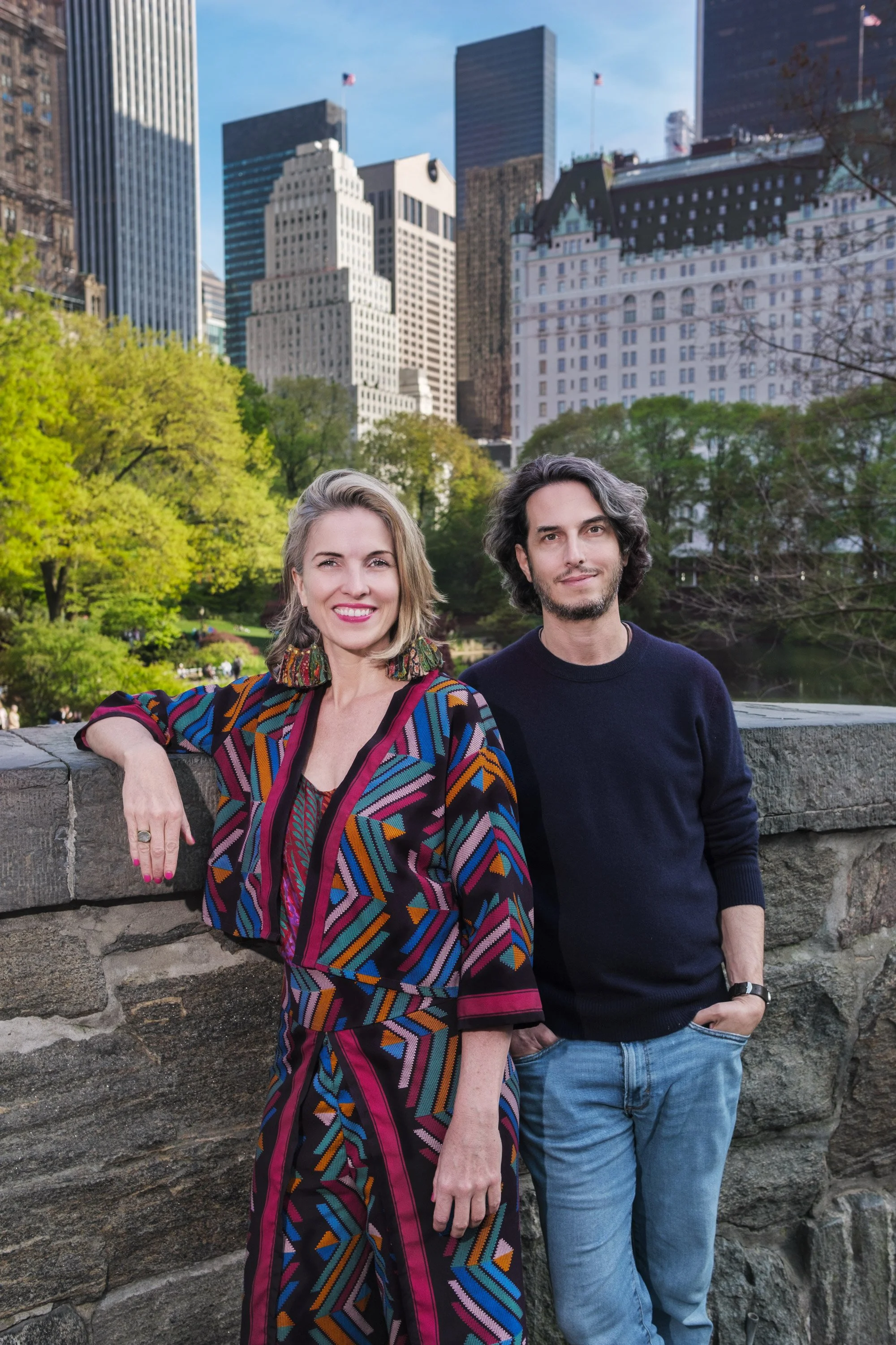 Two people, a woman with short blonde hair wearing a colorful, patterned jacket and a man with shoulder-length dark hair and a beard, wearing a dark sweater and jeans, standing next to a stone wall in a park with tall city buildings and trees in the background.