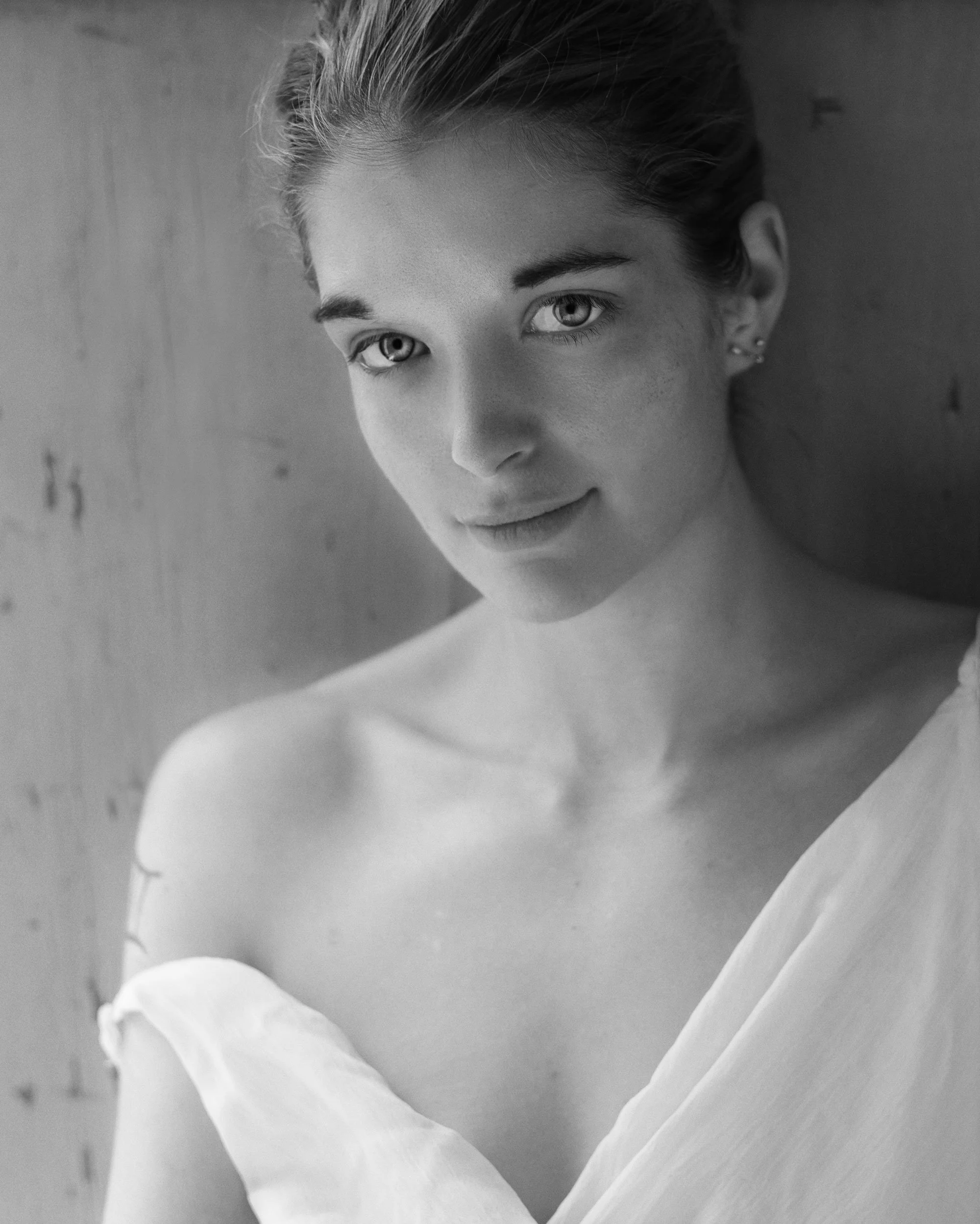 A close-up black and white portrait of a young woman with striking eyes, slicked-back hair, and a calm expression, partially viewed through a circular object.