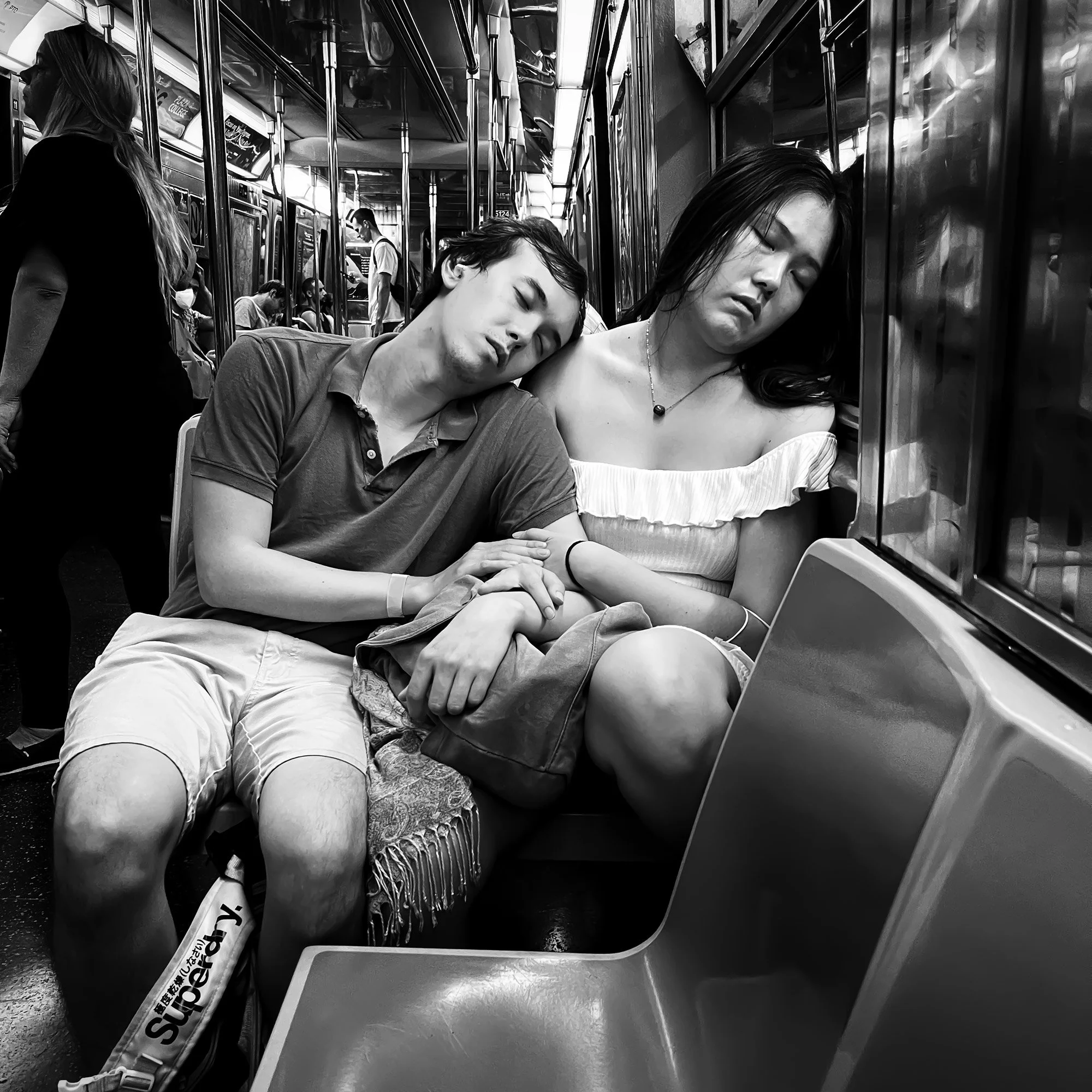 A black and white photo of a subway train interior with passengers. Two individuals are asleep with heads leaned against the window, one with short hair wearing a casual shirt, the other with long hair wearing a strapless top. Other passengers are visible in the background, some sitting, some standing, and a person riding a bicycle in the adjacent car.