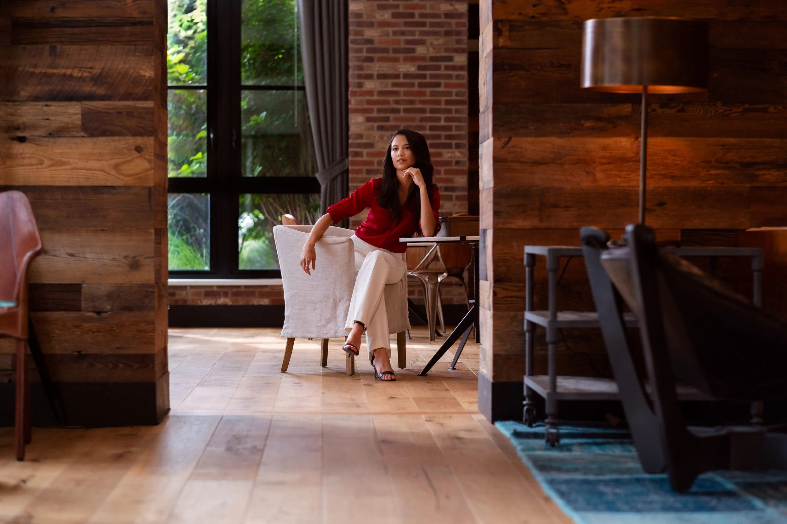 A woman with long dark hair sitting on a white chair inside a room with brick walls and large windows showing greenery outside. She is wearing a red top and white pants, with her right hand resting on her chin.