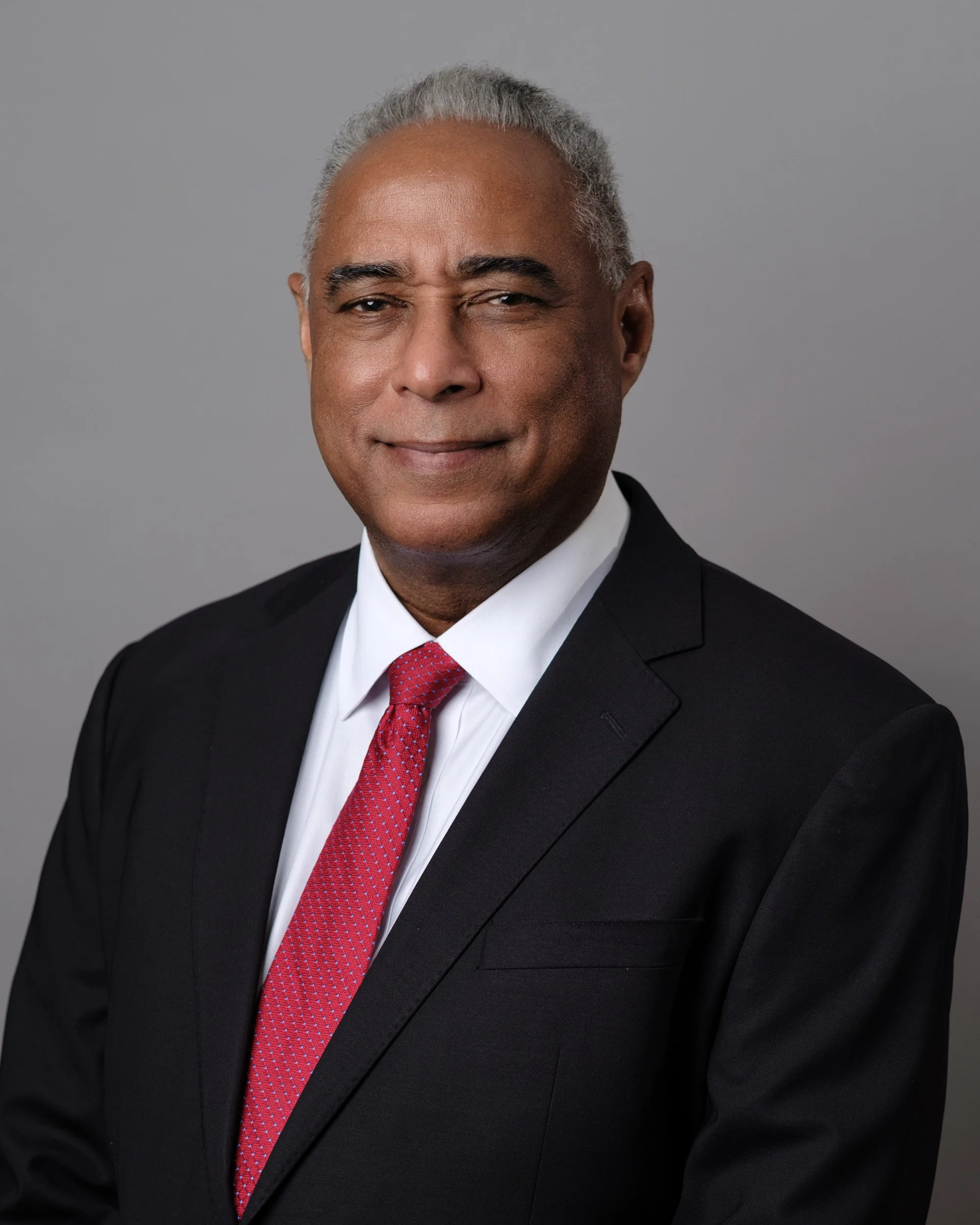 Close-up portrait of a middle-aged African American man in a black suit, white shirt, and red tie, smiling with one eye slightly squinted against a plain gray background.