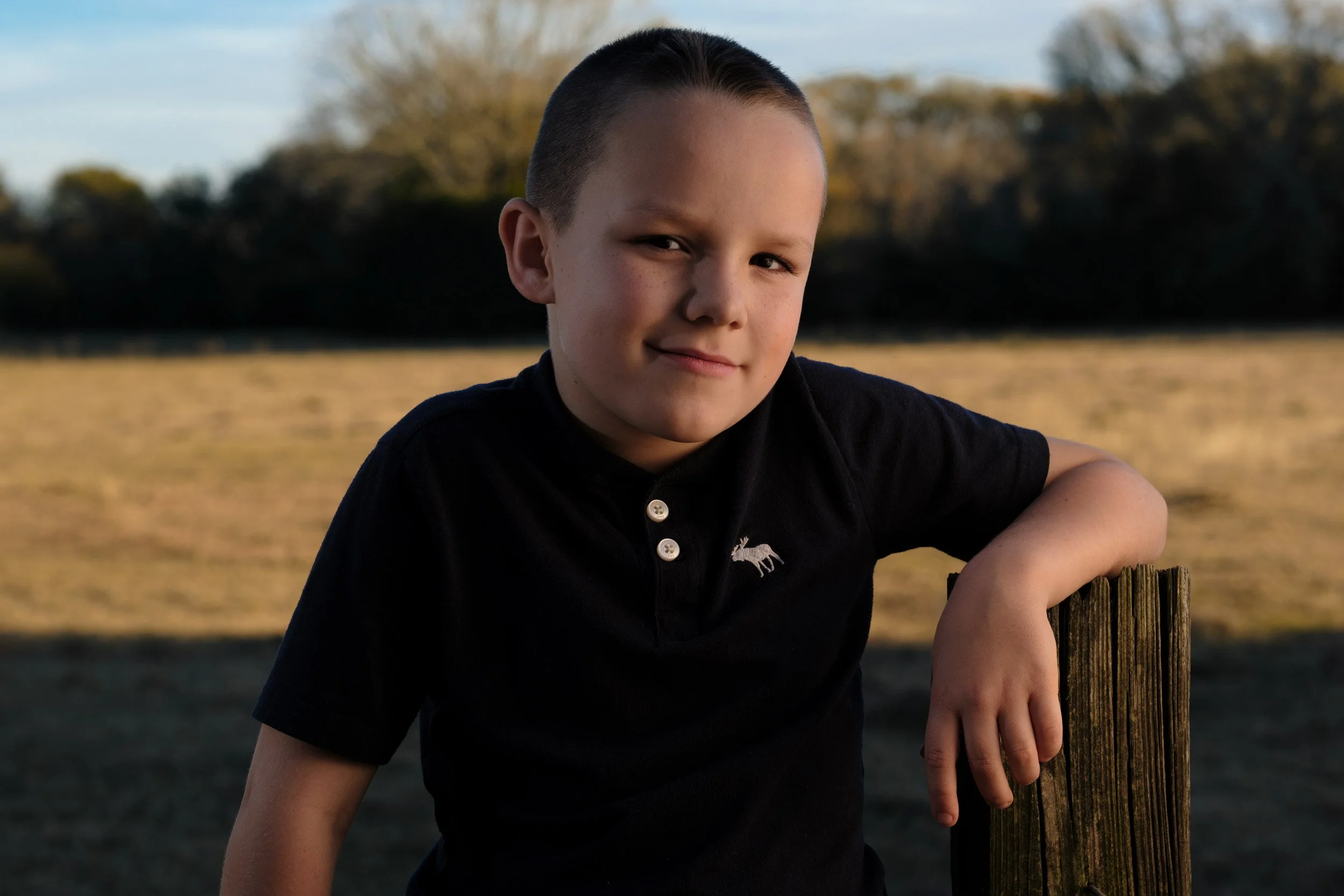 A young boy with short hair wearing a black polo shirt with a moose logo, standing outdoors during late afternoon, leaning on a wooden post with a blurred background of trees and grass.