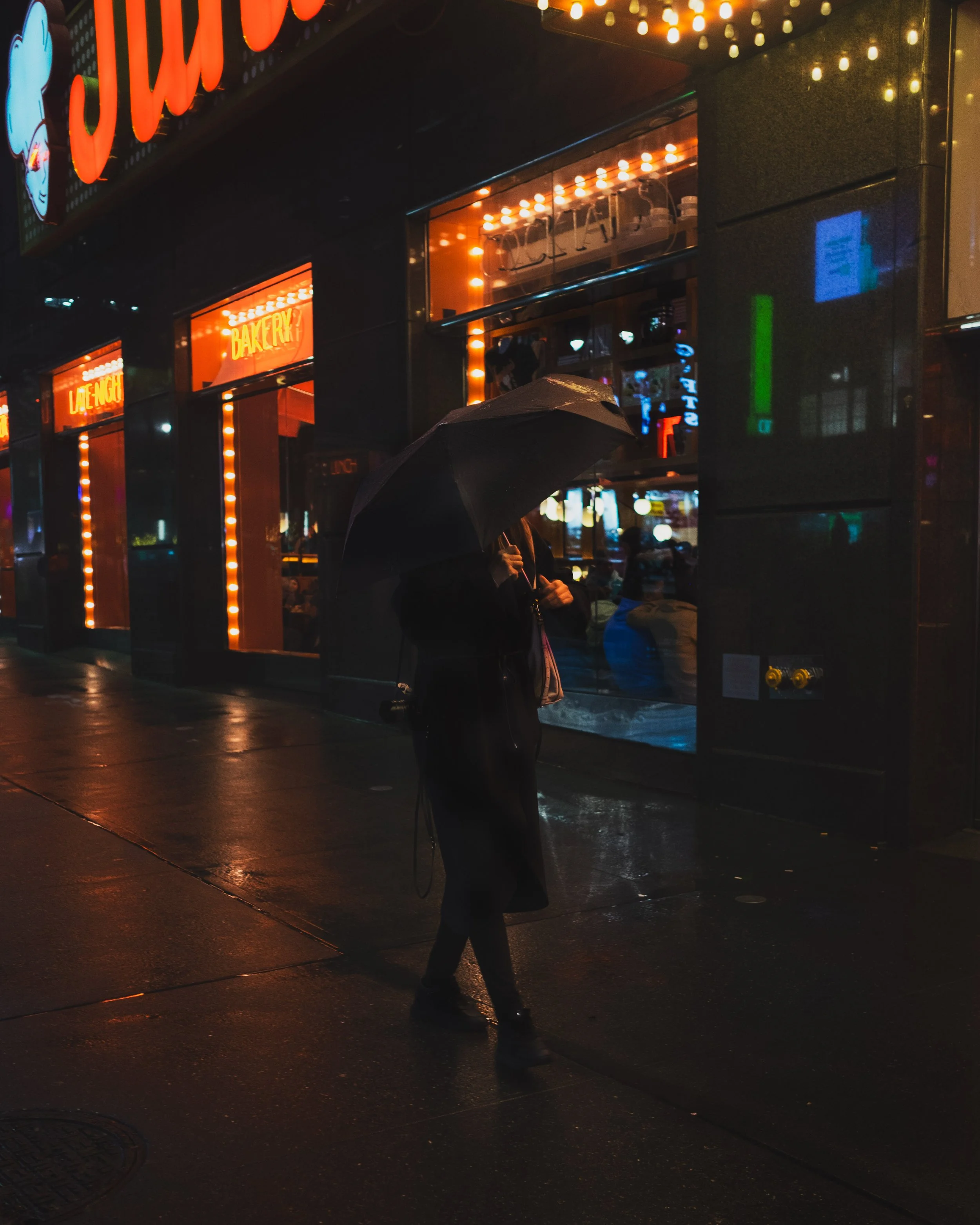 A person holding an umbrella walking on a wet sidewalk at night in front of a store with bright orange and yellow neon signs, glare reflections on the pavement, and a dark rain-soaked street.
