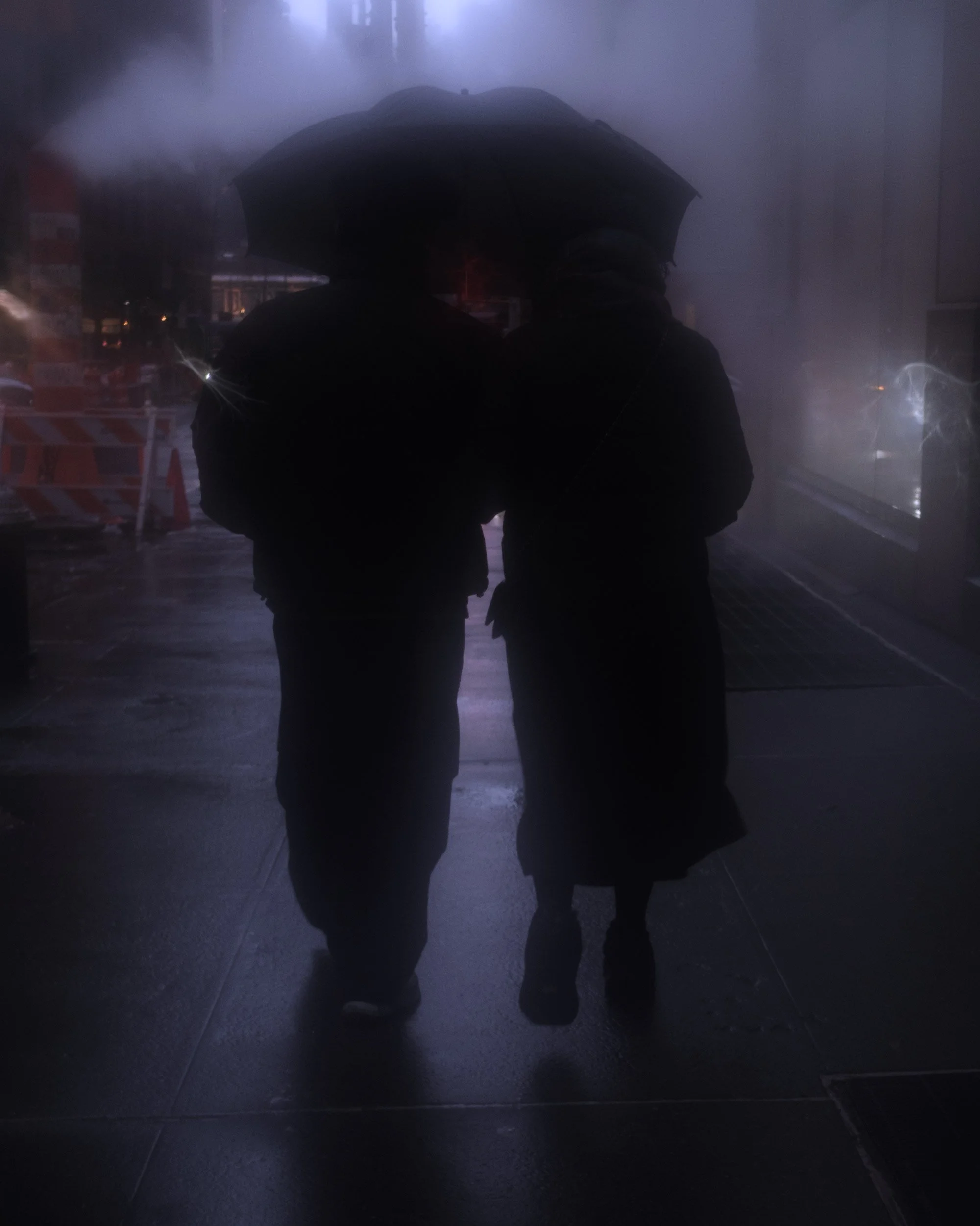 Two individuals standing close together under an umbrella during a rainstorm, silhouetted against a reflective, wet sidewalk in an urban setting at night.
