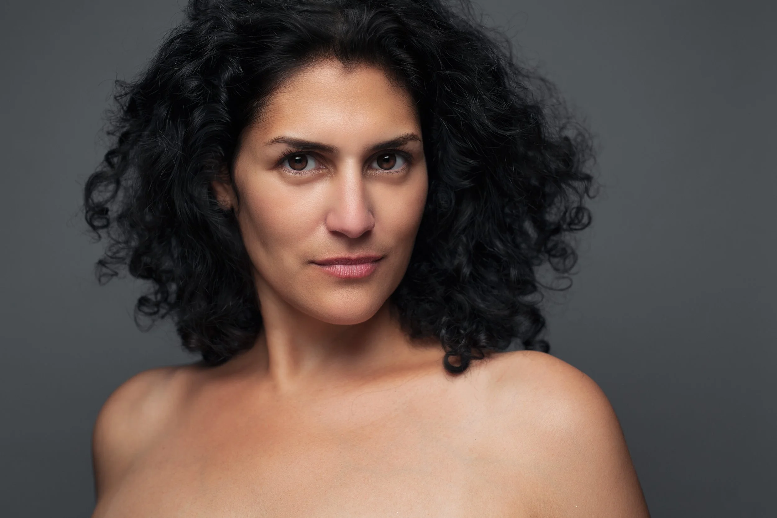 Close-up portrait of a woman with curly black hair and brown eyes, against a gray background, with bare shoulders and a subtle smile.