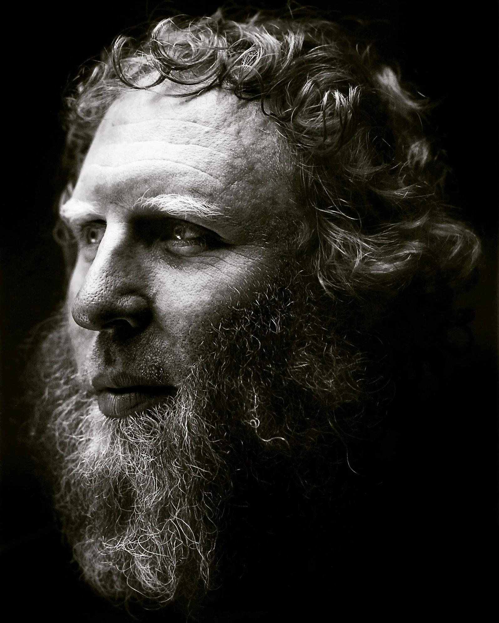 Close-up black and white photograph of an elderly man showing his face in profile. He has curly hair, a beard, and visible wrinkles.