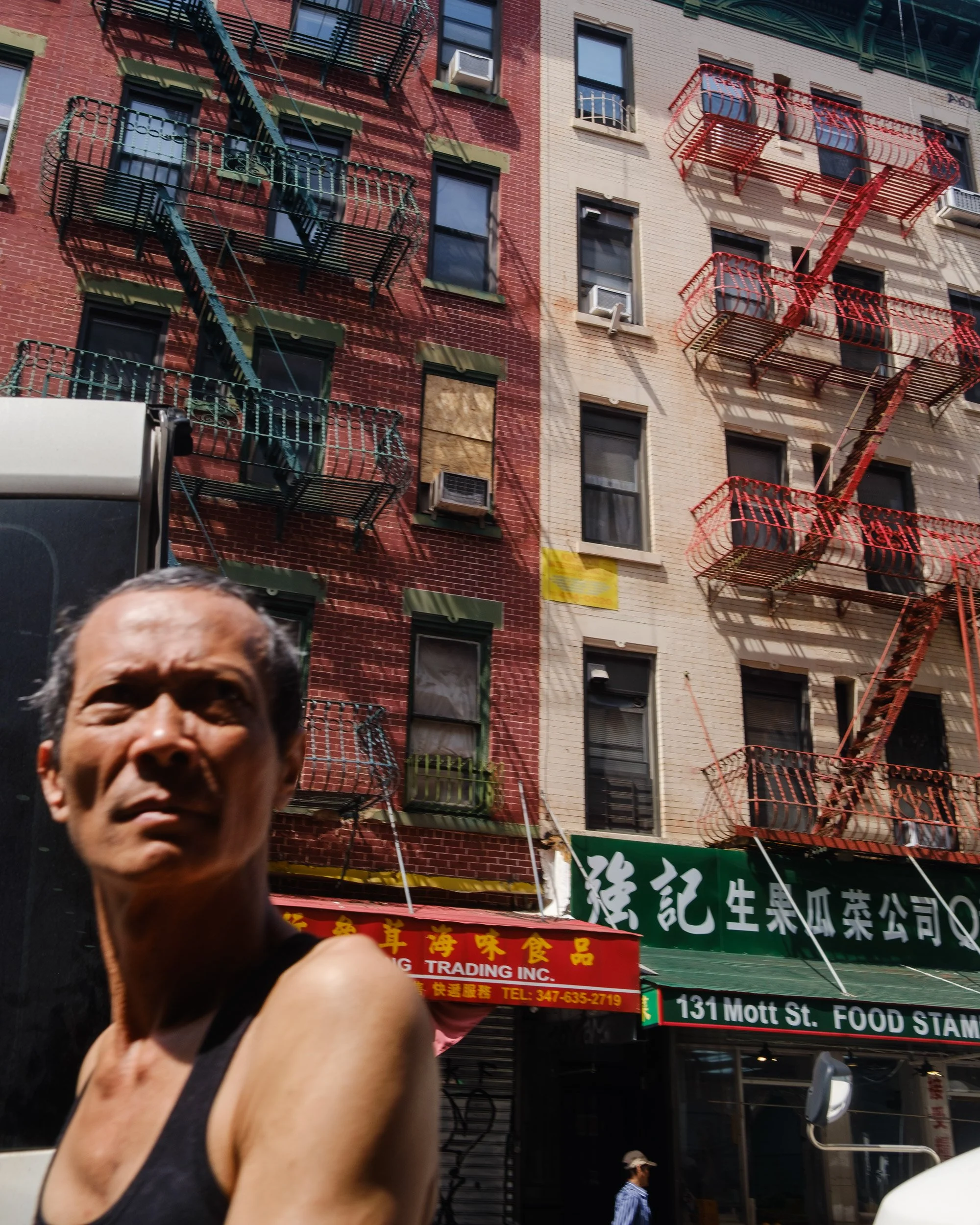 A man smiling and giving a thumbs-up in front of a multistory building with fire escapes and Chinese signage along a street.