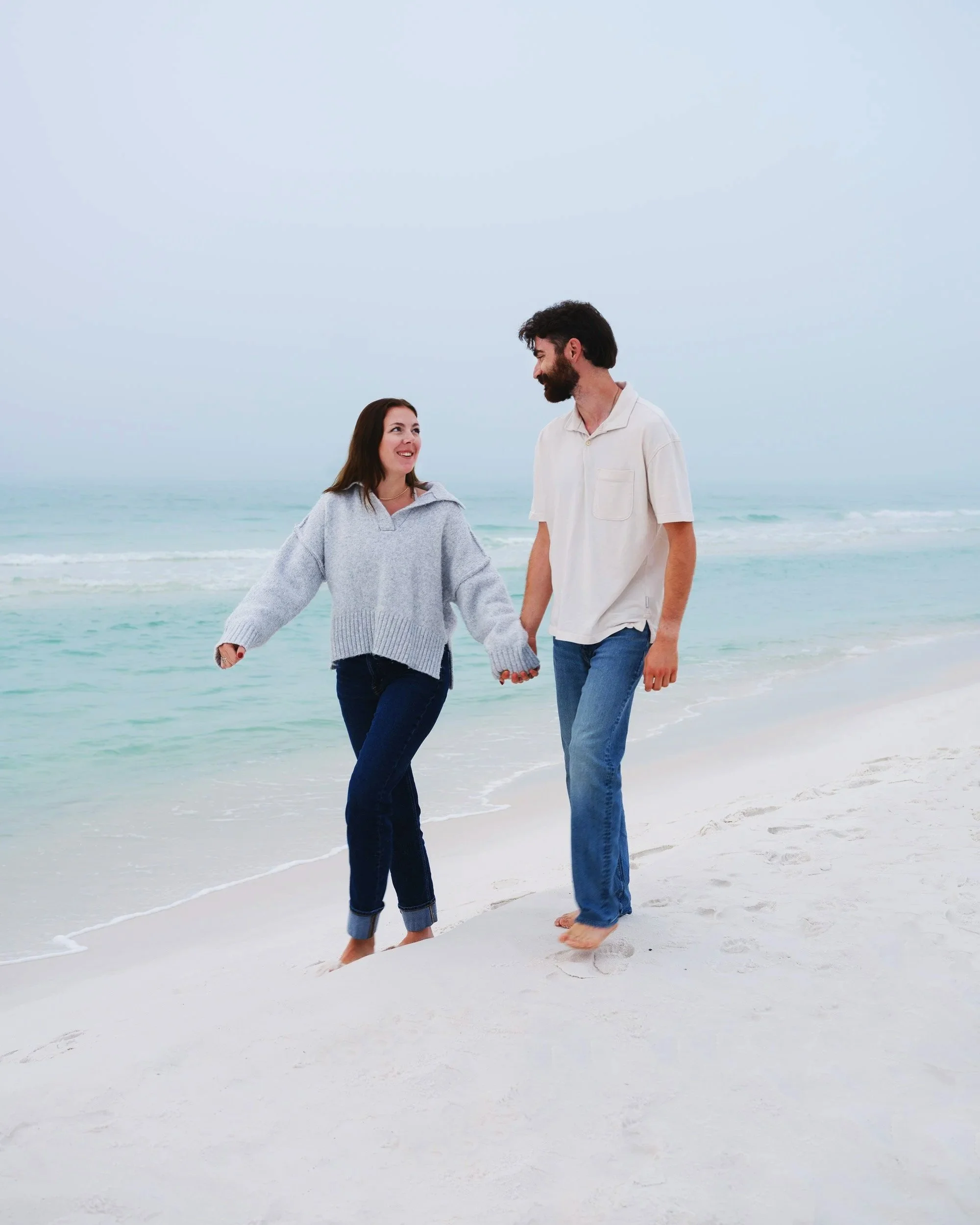 A smiling couple holding hands and walking on a sandy beach near the ocean, wearing casual clothes, with the woman wearing a grey sweater and the man wearing a white shirt.