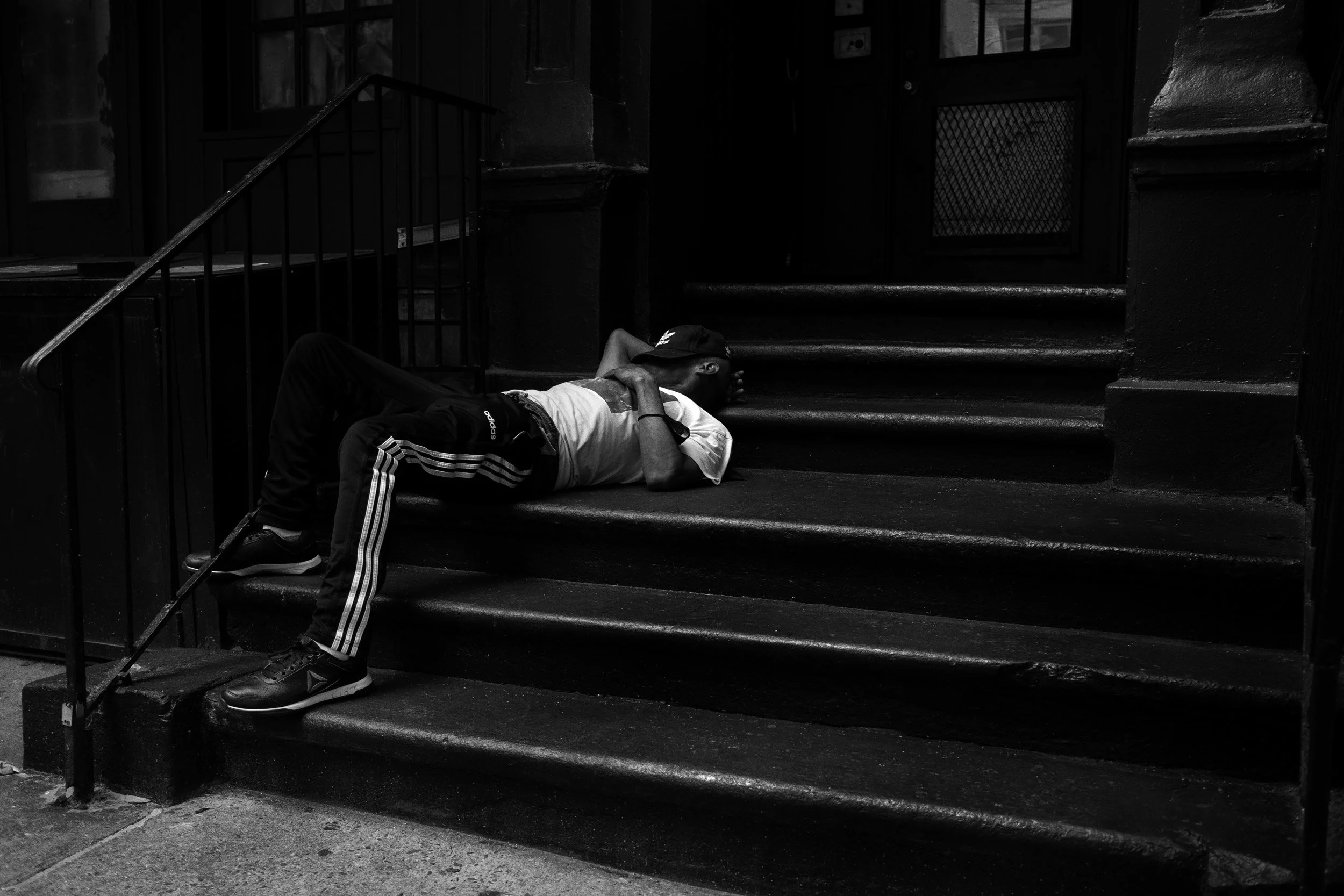 A person lying on a staircase in an urban setting, wearing a cap, t-shirt, and Adidas pants, with a dark environment surrounding them.