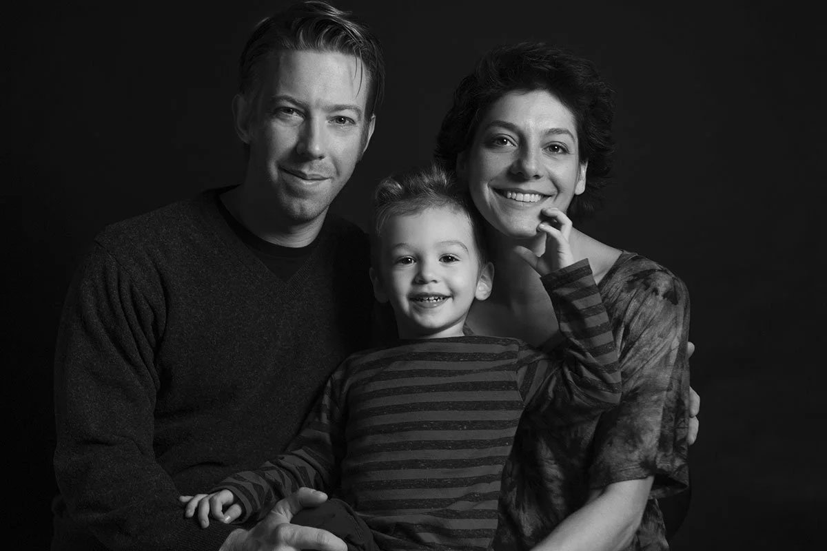 Black and white photo of a smiling family with a man, woman, and a young girl with curly hair, all sitting close together against a plain dark background.
