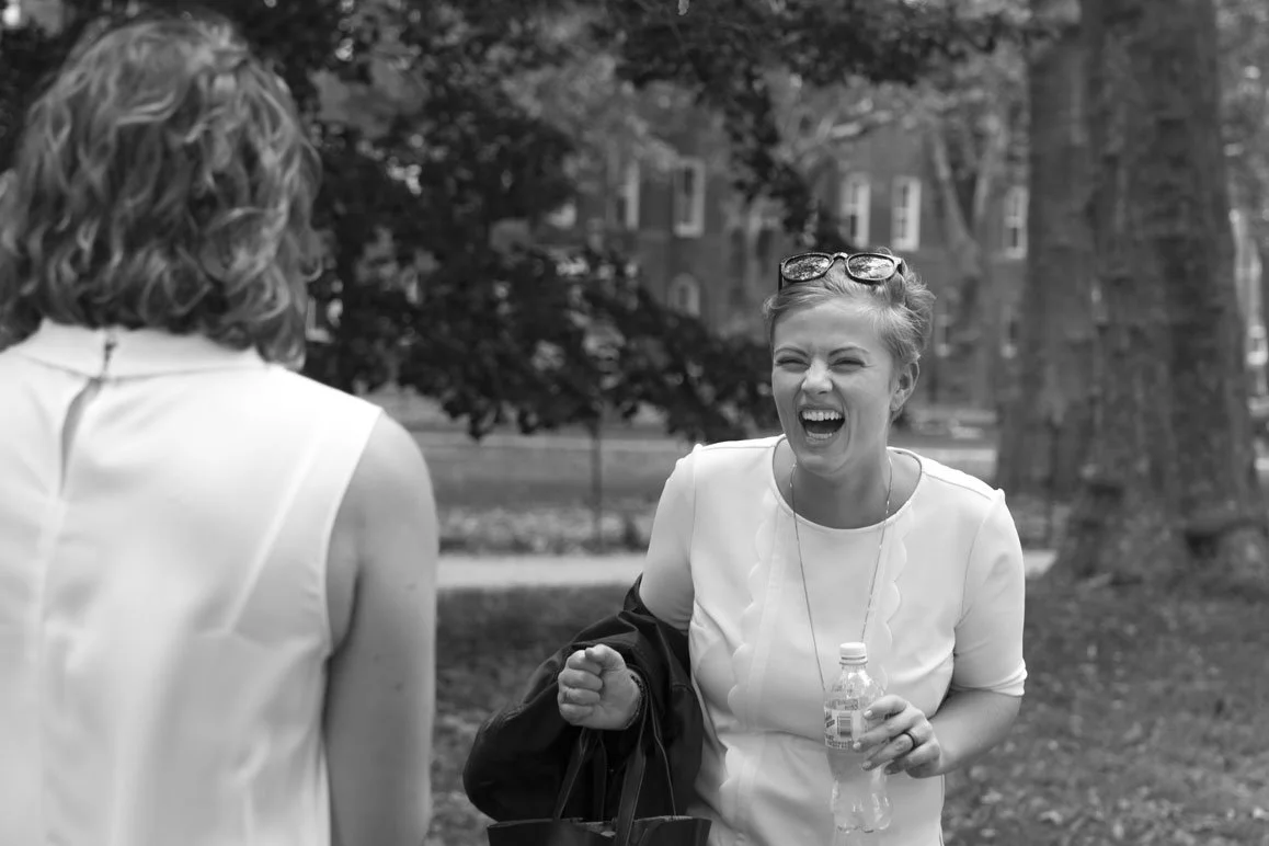 Two women laughing and talking outdoors in a park, one holding a water bottle, with trees and buildings in the background.