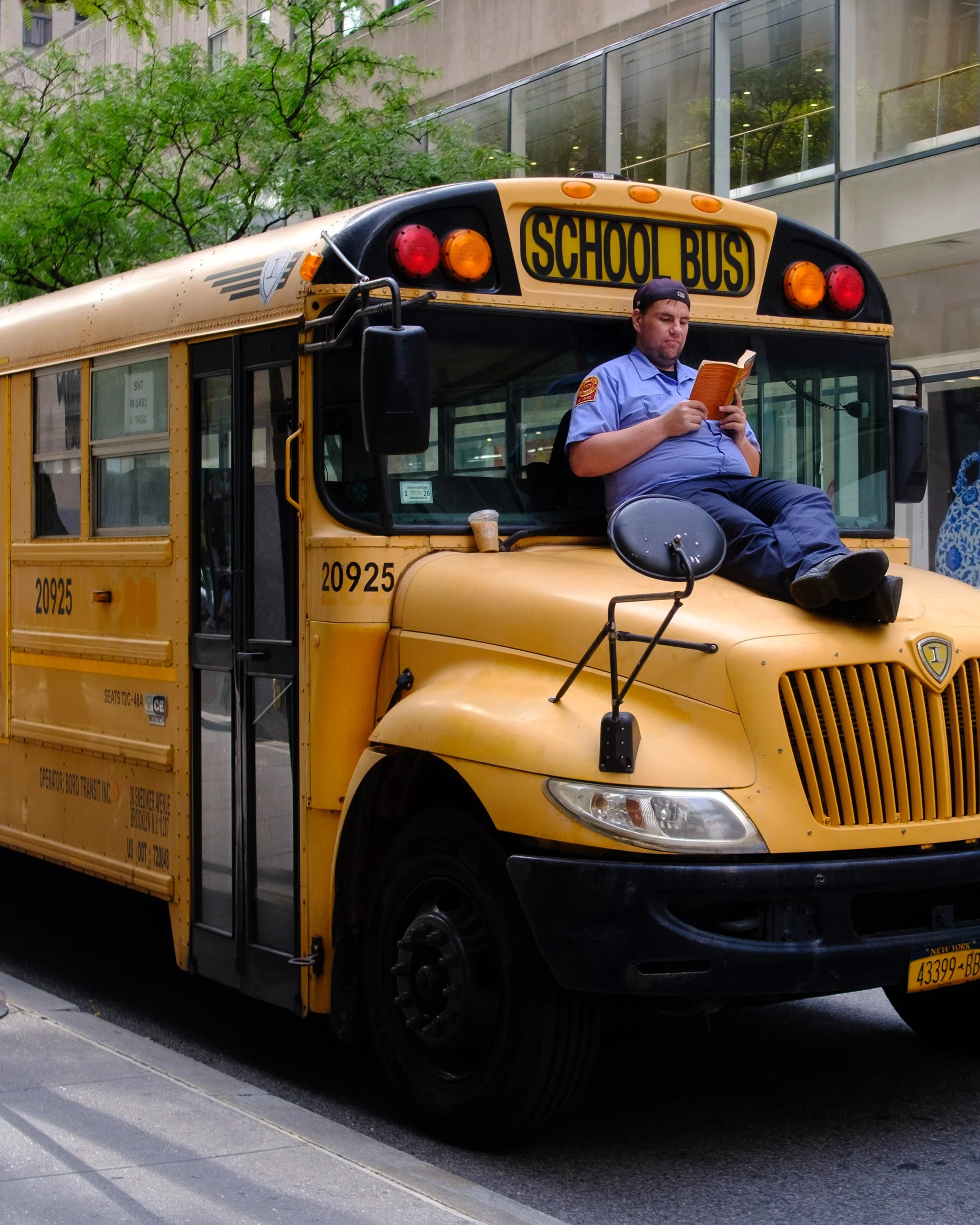 A yellow school bus parked on the side of a city street with a traffic cone on its front bumper. A city building with glass windows and trees in the background. A man in a blue uniform and a black cap is sitting on the bus's front bumper, reading a b