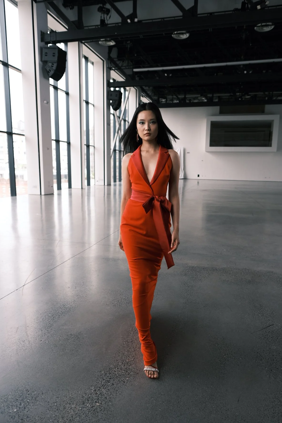 A woman in an orange sleeveless dress with a deep V neckline and a tied waist walks in an empty modern building with large windows and high ceilings.