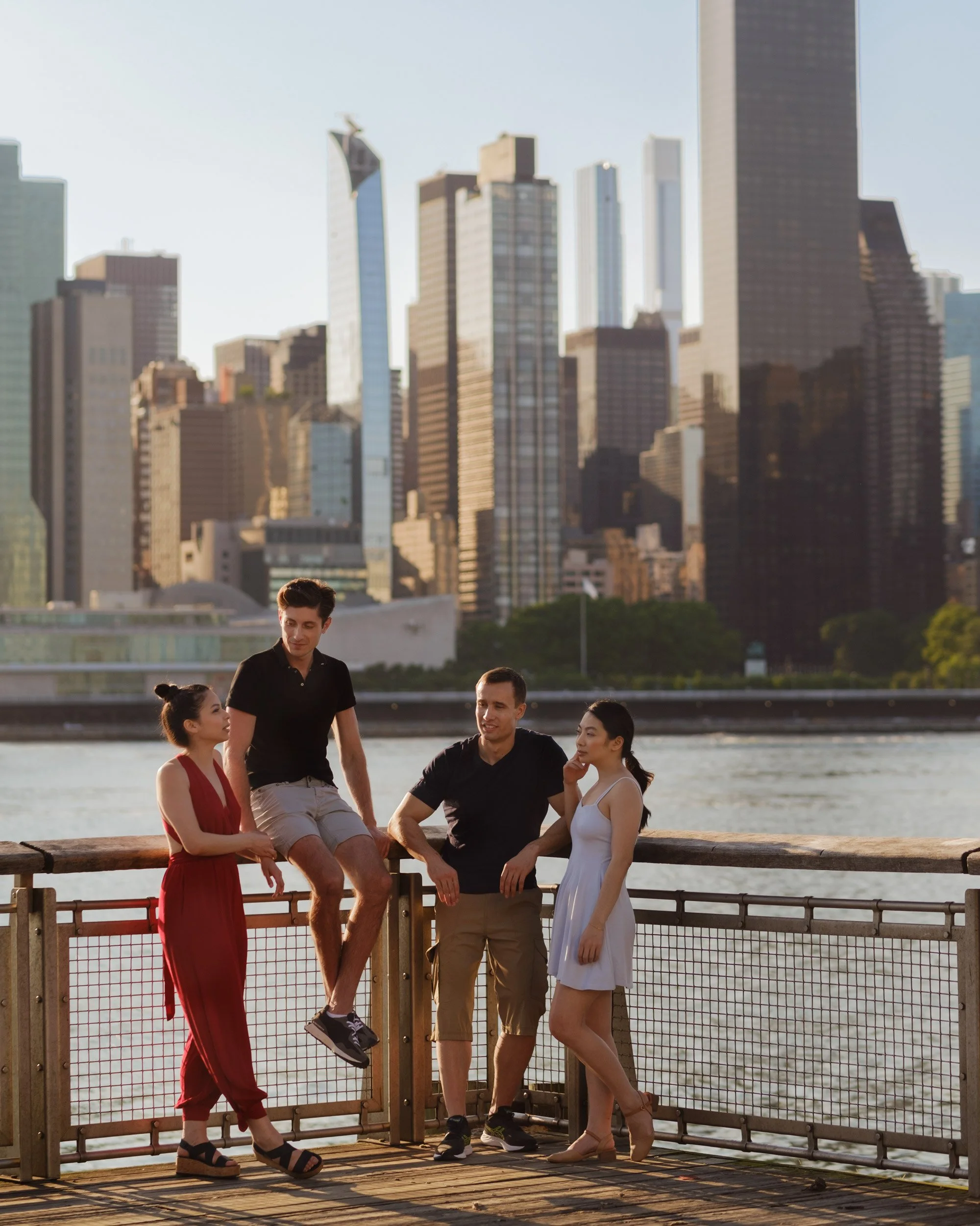 Five young adults are gathered on a wooden dock by the water, engaging in conversation. Behind them is a city skyline with tall buildings and skyscrapers. The scene is outdoors during sunset or late afternoon.
