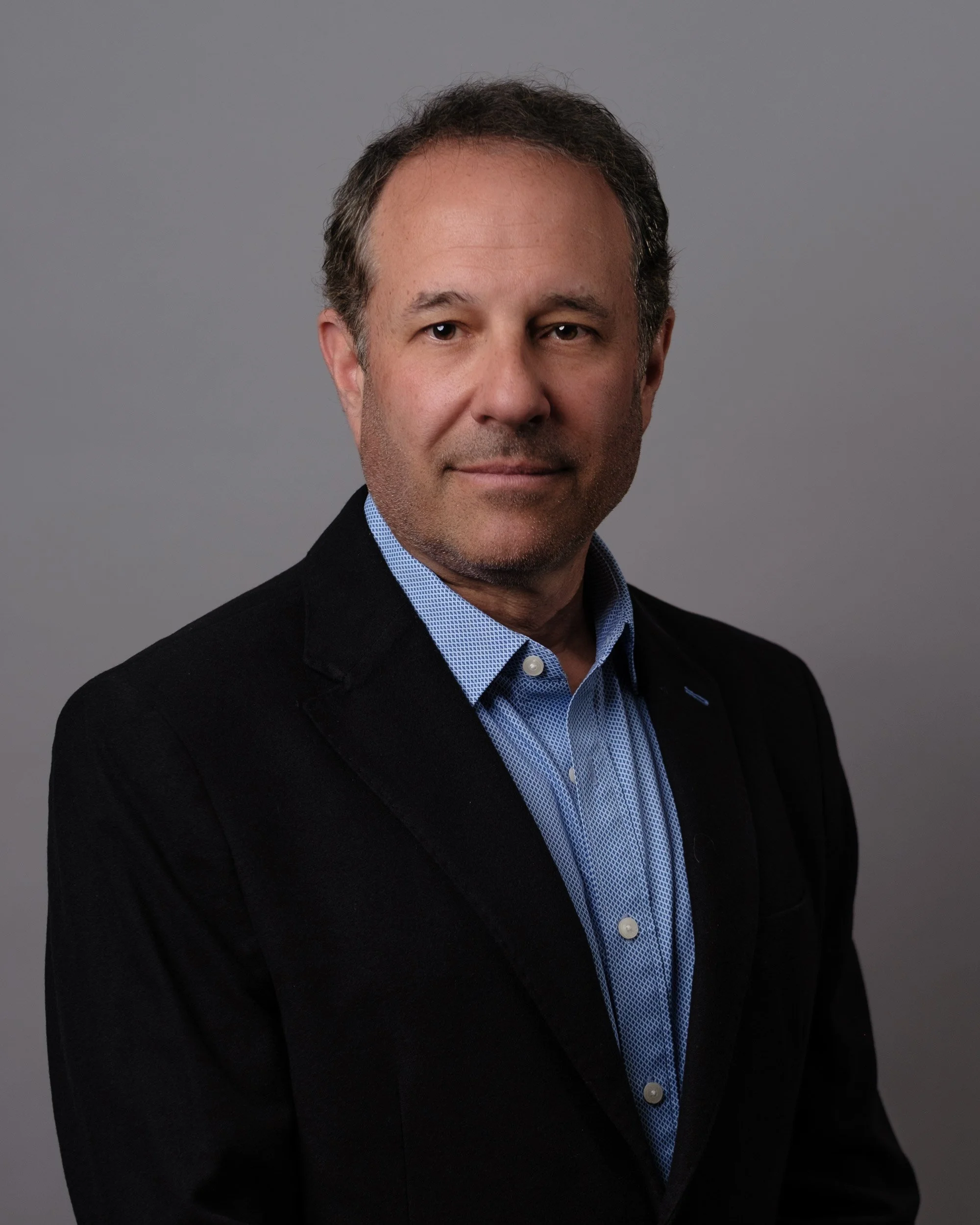 A headshot of a middle-aged man with dark hair, wearing a blue collared shirt and black blazer, against a gray background.
