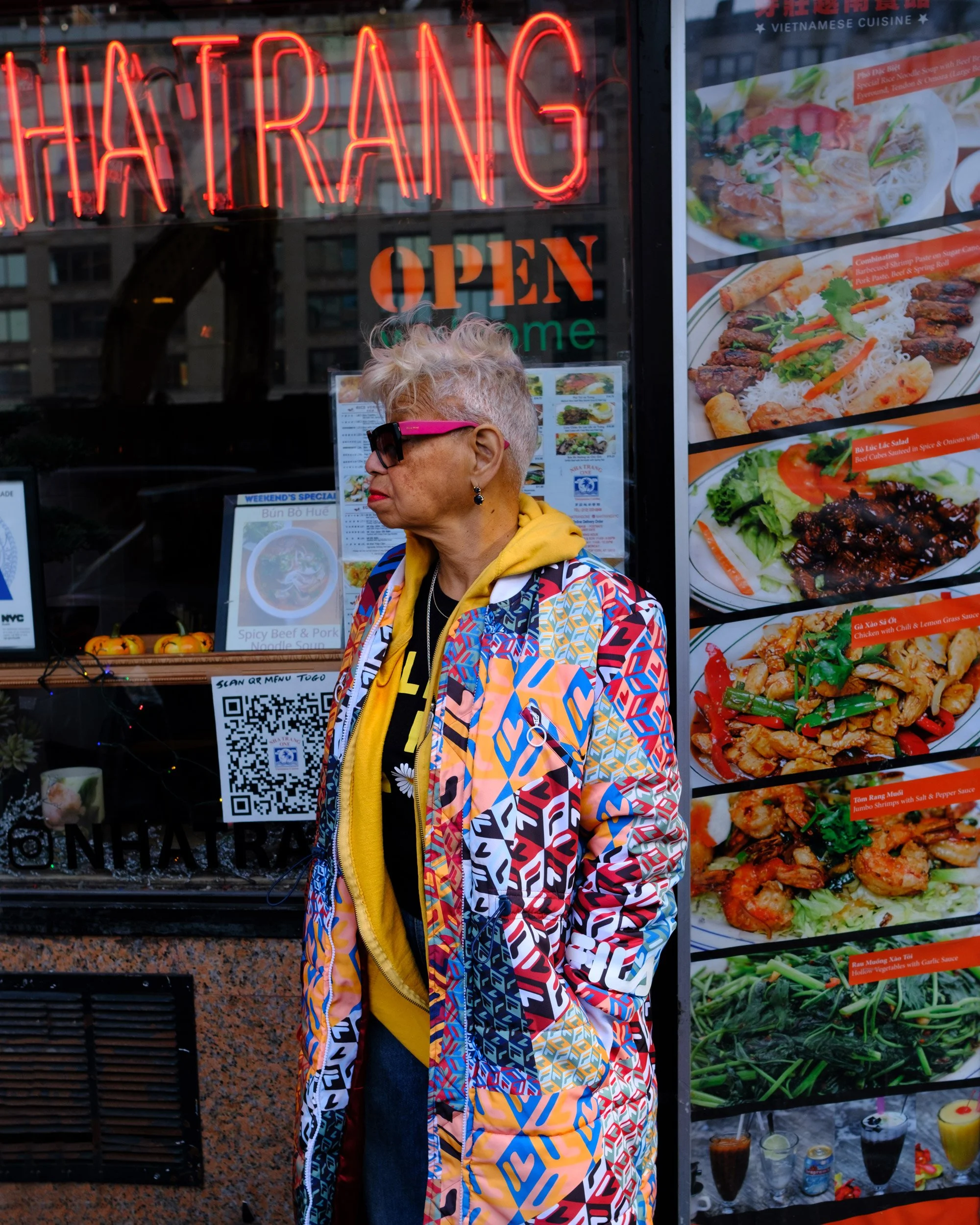 A woman with brightly dyed blonde hair, wearing sunglasses, a yellow hoodie, and a colorful patterned jacket, standing outside a Vietnamese restaurant with a neon 'HÁI TRANG' sign and menu images of Asian dishes displayed on the window.