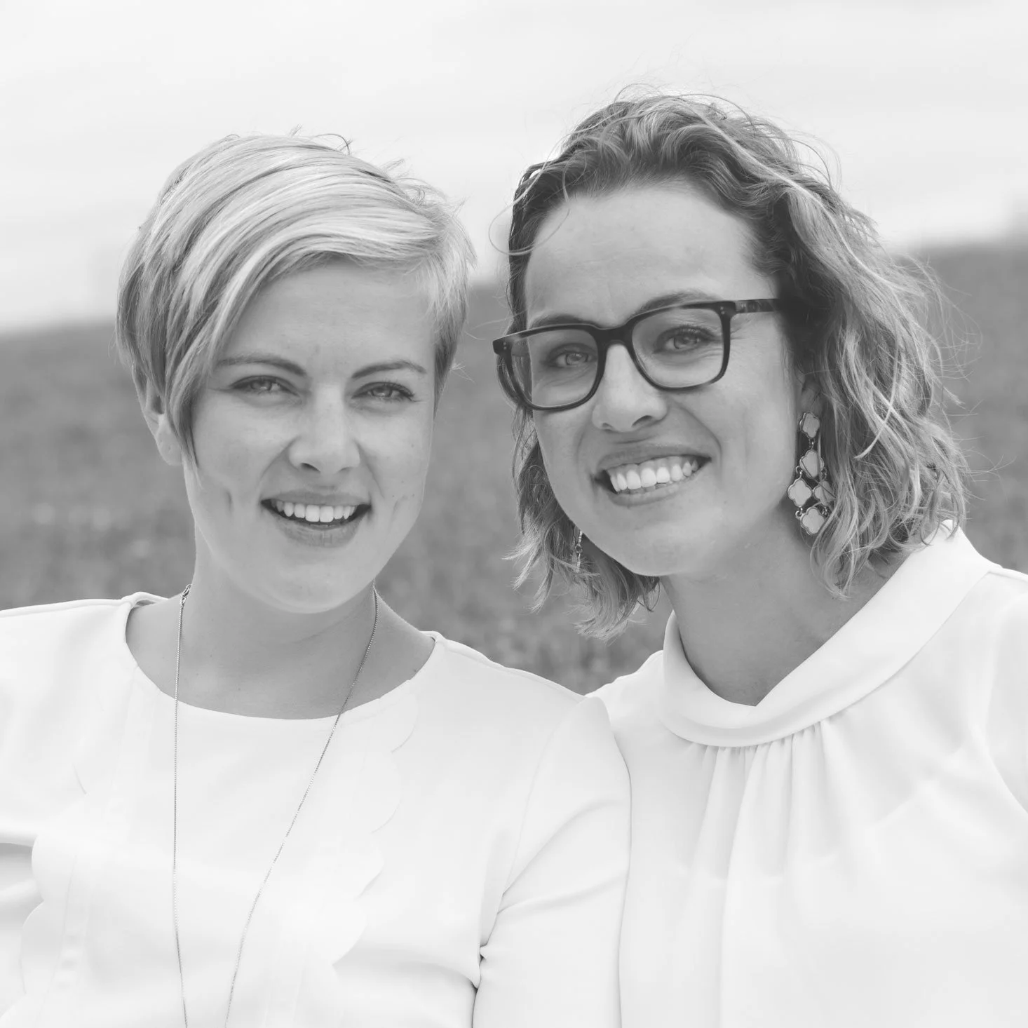 Two women smiling outdoors in black and white, standing close together with their heads touching. The woman on the left has short, light-colored hair, and the woman on the right has wavy hair, wears glasses, and large earrings.