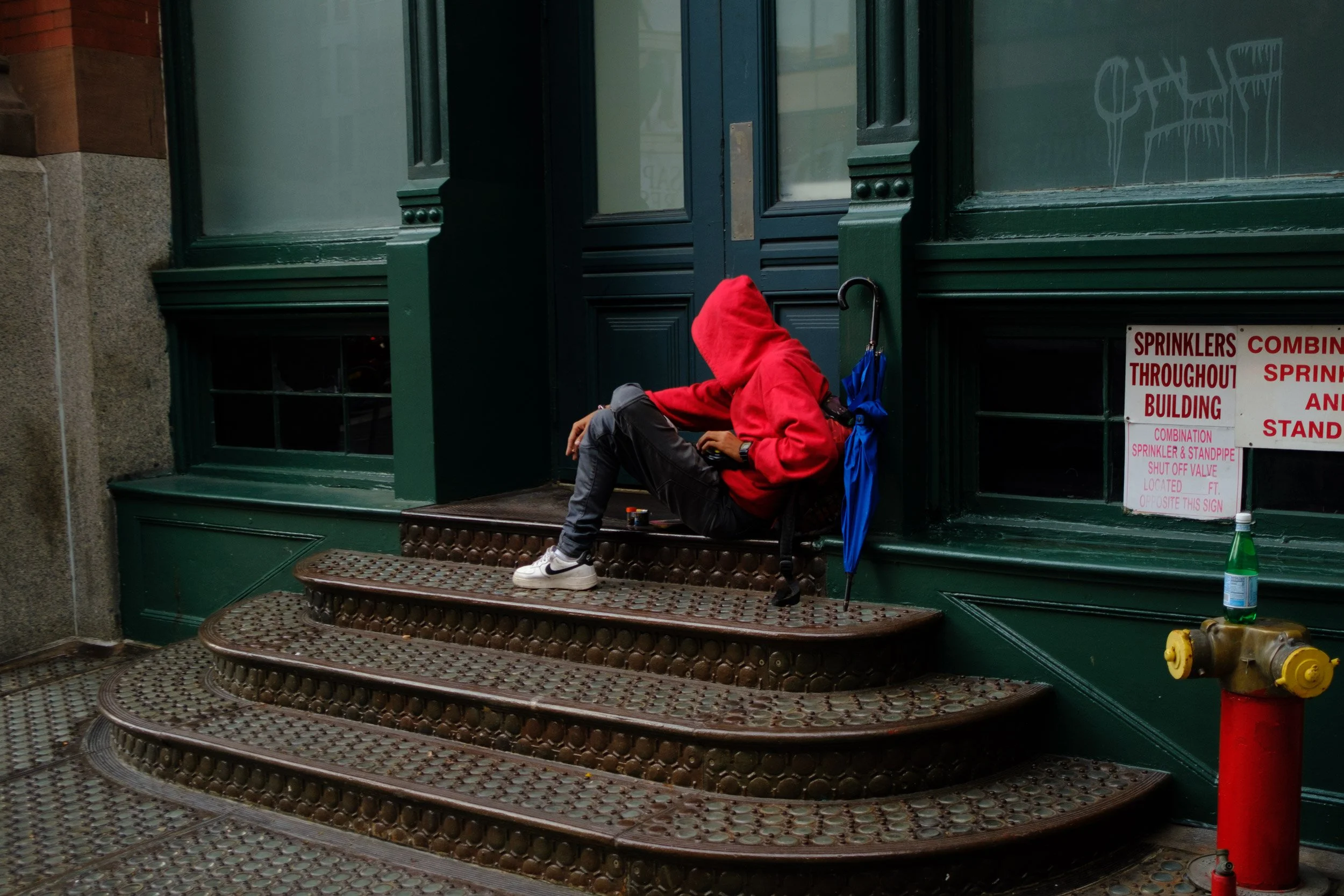 A person wearing a red hoodie and black pants sitting on a patterned metal staircase outside a green storefront, with a sign about sprinkler maintenance and a leaning blue umbrella next to them.