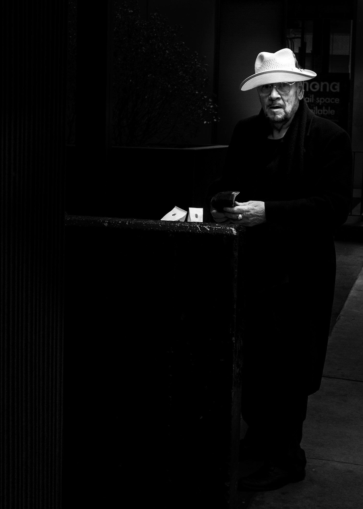 A black and white photo of an older man wearing glasses, a large sunhat, and a dark jacket. He is standing outdoors, holding a mobile phone in his right hand, looking towards the camera with a surprised or confused expression.