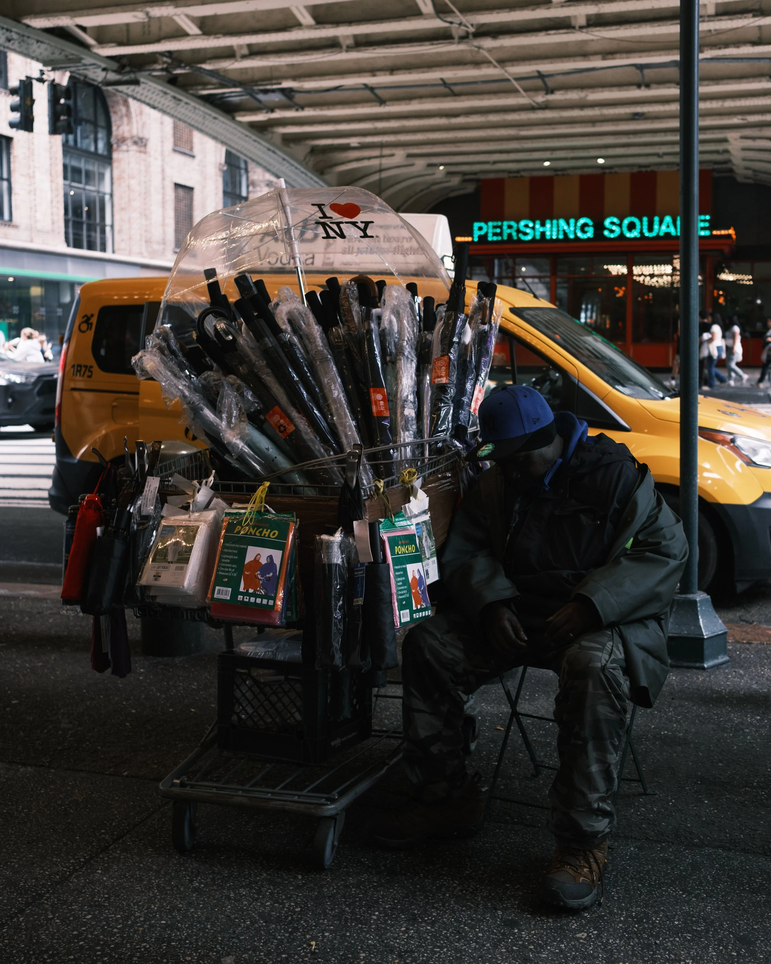 A street scene in New York City, featuring a man in camouflage pants and outdoors gear sitting on a folding chair beneath a covered sidewalk, with a cart full of umbrellas and packaged merchandise nearby. Yellow taxis and a storefront with a neon sig