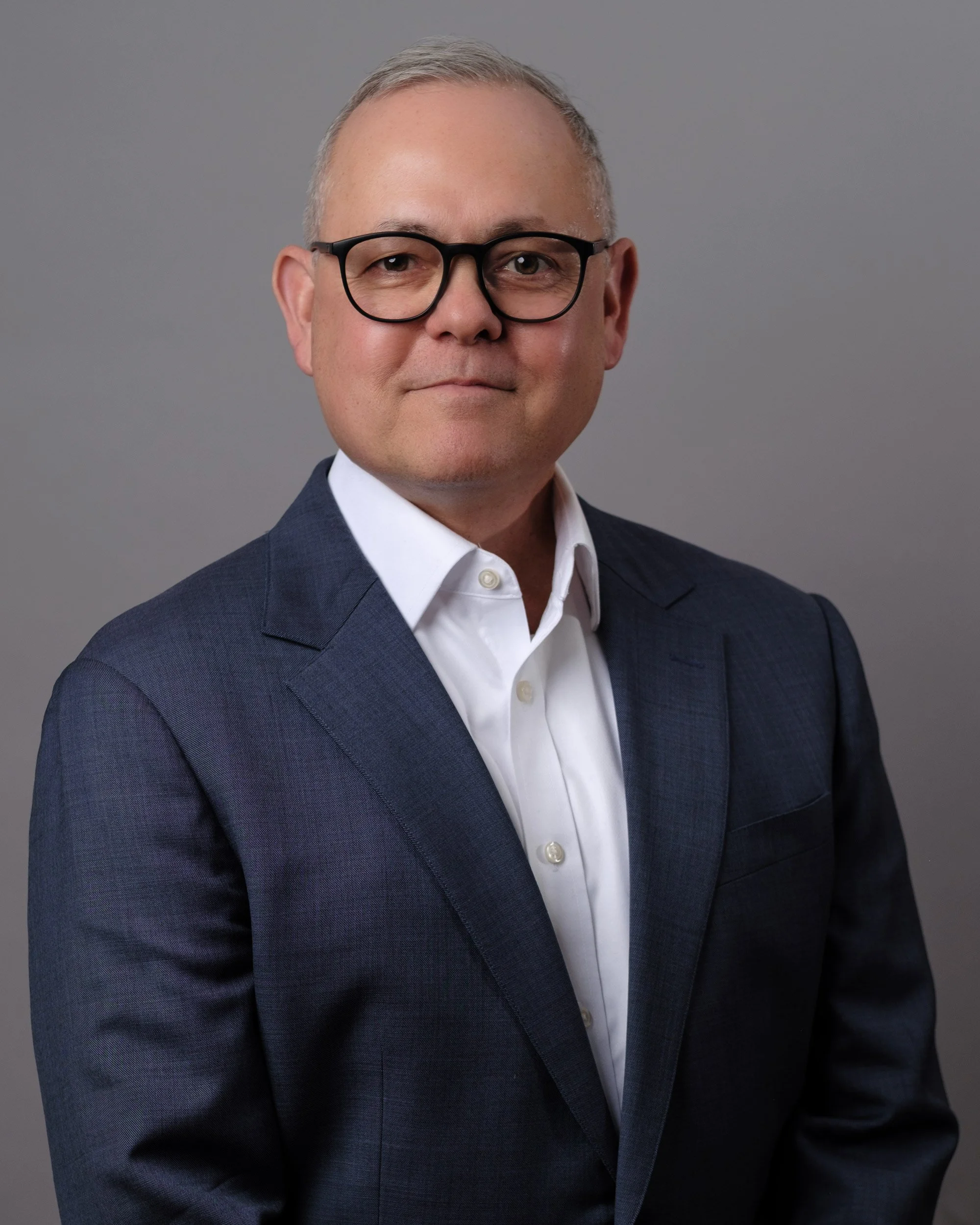 A middle-aged man with short gray hair, glasses, wearing a dark blue suit and white shirt, posing against a plain gray background.