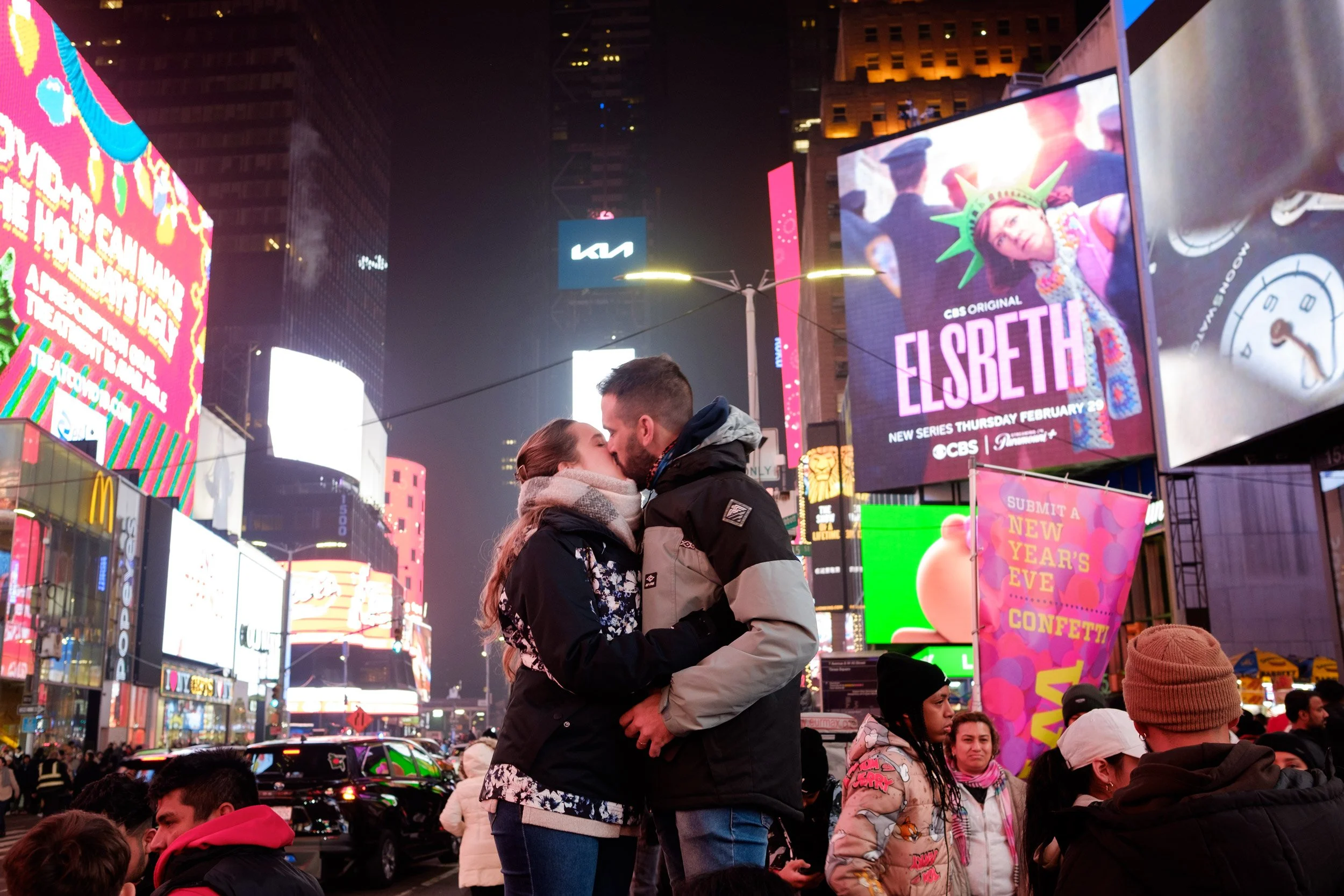 A couple sharing a tender moment in Times Square at night, surrounded by illuminated billboards and a crowd of people.