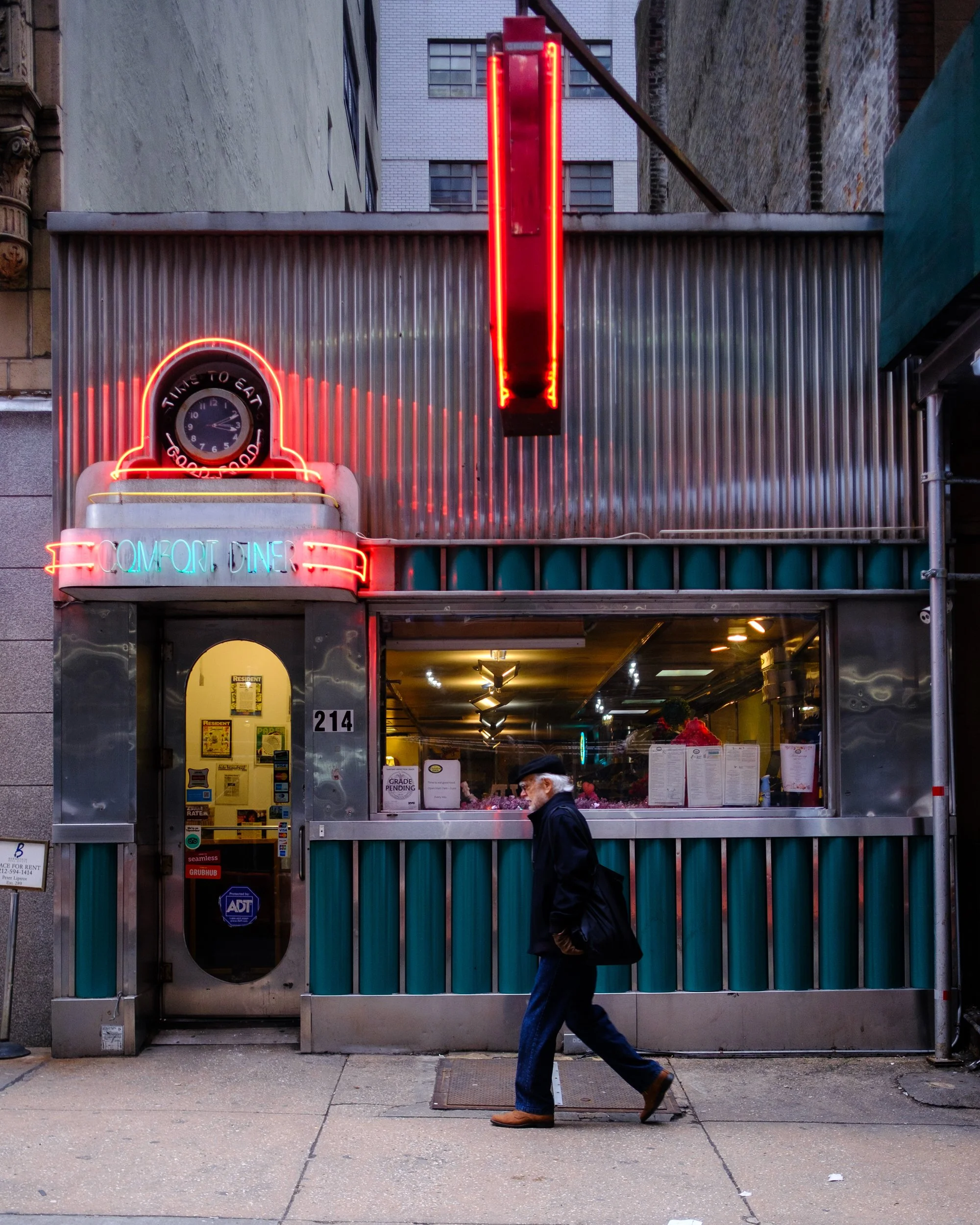 A person dressed in dark clothing and wearing brown shoes walks past a diner with neon signs that read 'Comfort Diner'. There is a large neon red vertical sign hanging above the diner entrance, and a neon clock that says 'Time to Eat'. The diner's ex