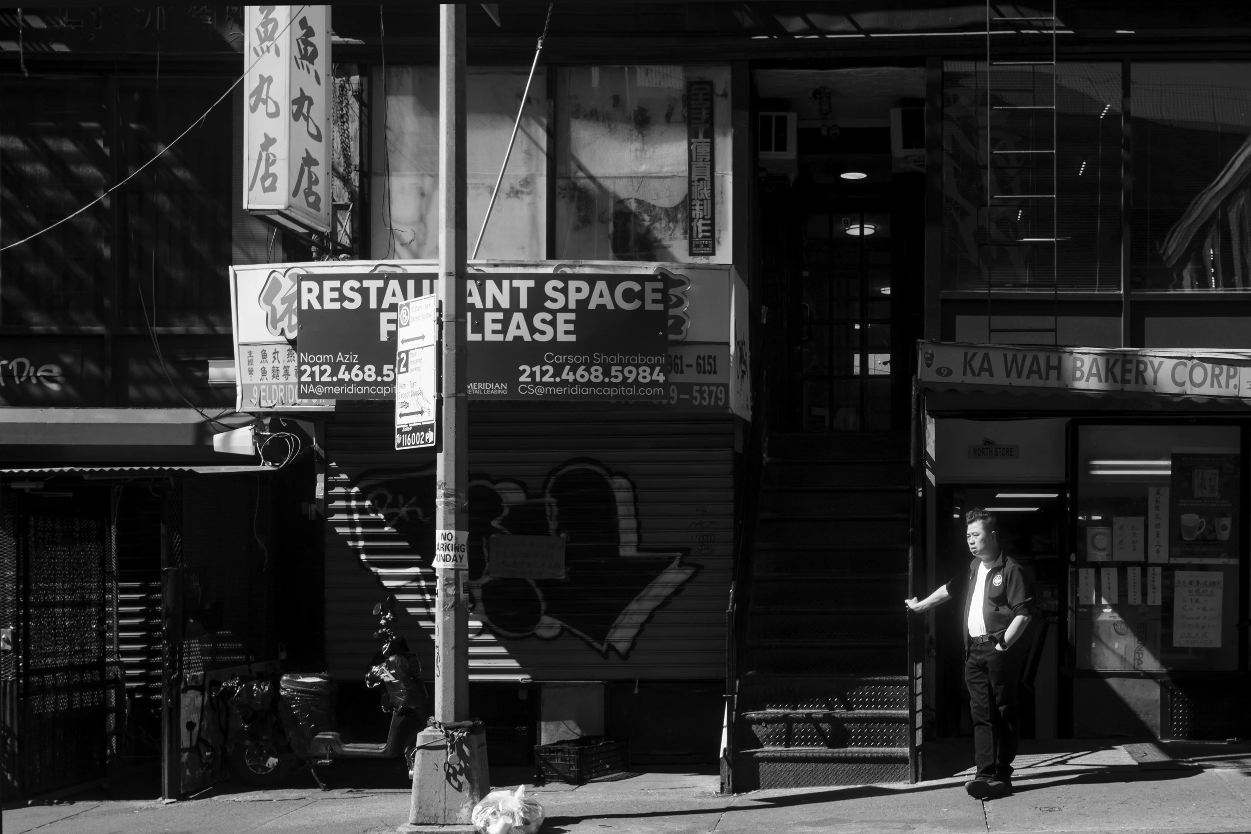 Urban street scene in black and white, with storefronts and signage, a person standing with hands in pockets, and graffiti on a closed shop shutter.